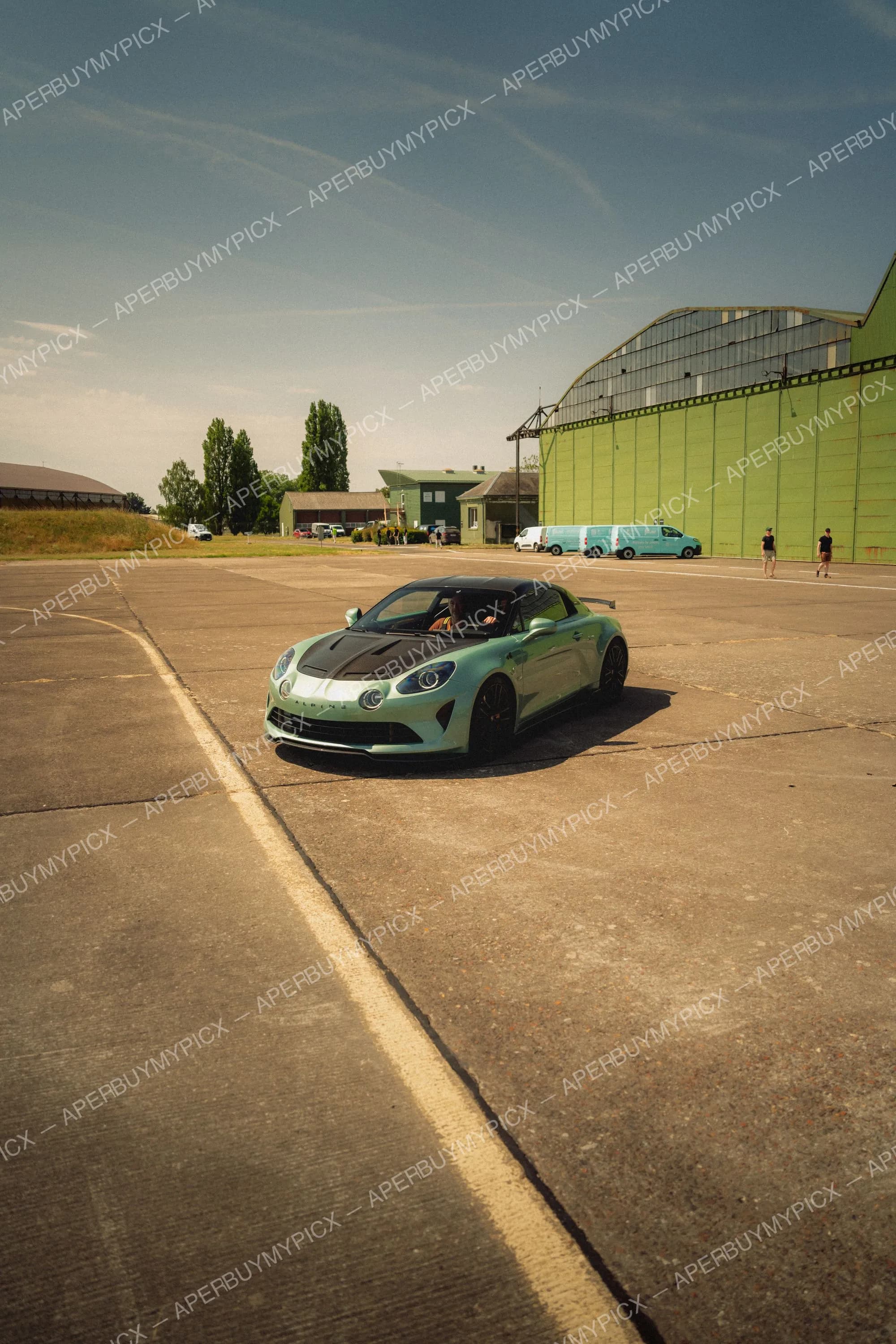 Alpine A110 GT vert menthe avec capot en carbone garée sur un tarmac d'aérodrome, devant un hangar vert, sous un ciel bleu ensoleillé.