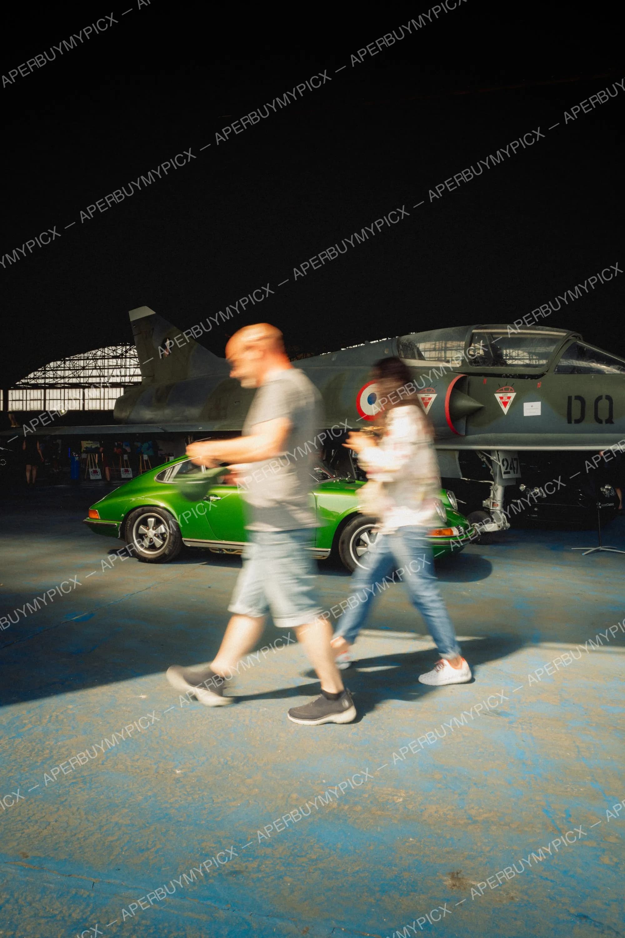 Une Porsche 911 verte vintage stationnée devant un avion de chasse Mirage vert militaire dans un hangar, tandis qu'un couple flou marche au premier plan sur un sol bleu.