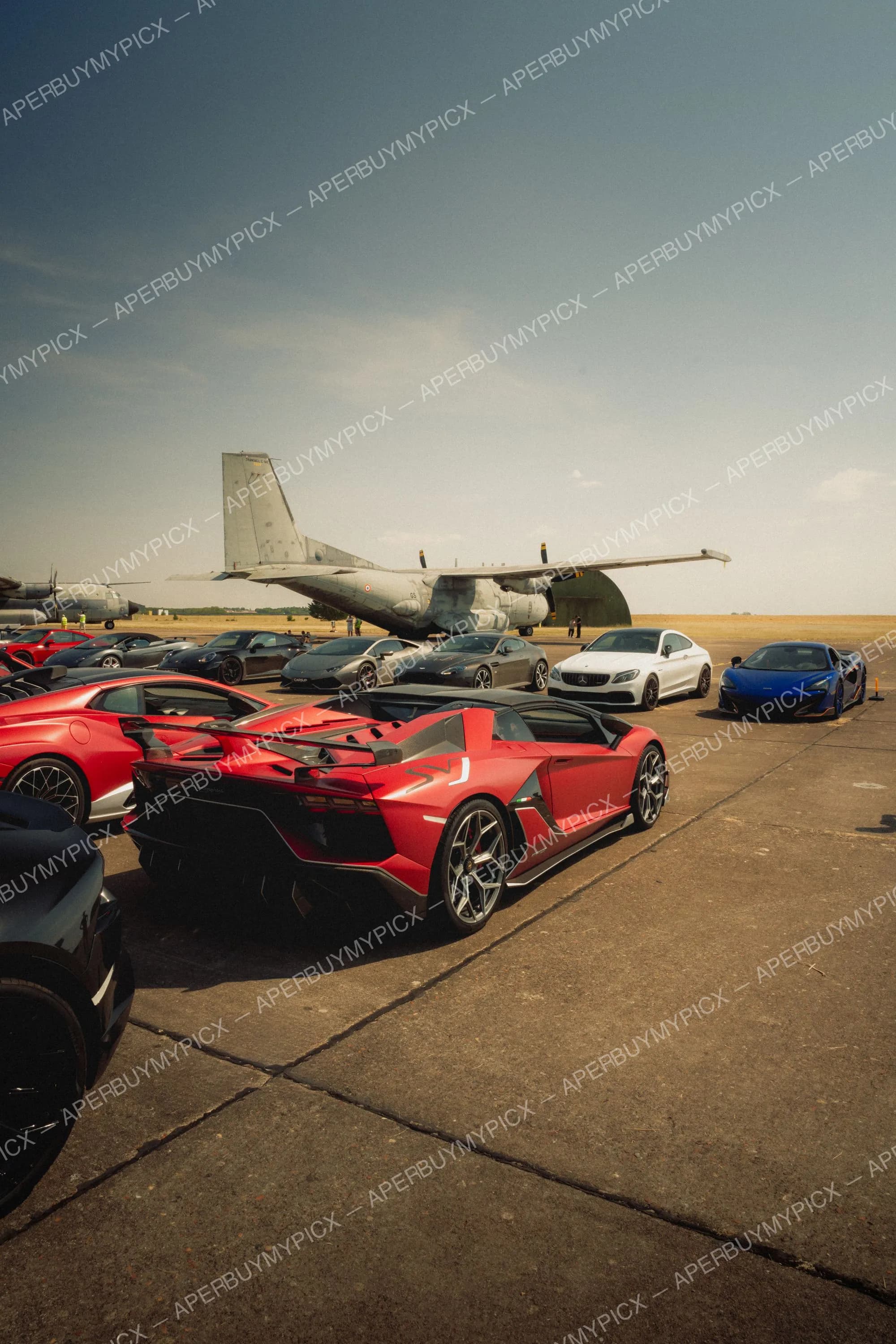 Lamborghini Aventador SVJ rouge décapotable garée sur le tarmac d'une base aérienne, entourée de supercars dont une McLaren bleue, une Mercedes-AMG blanche et des Porsche, devant un avion de transport militaire sous un ciel dégagé