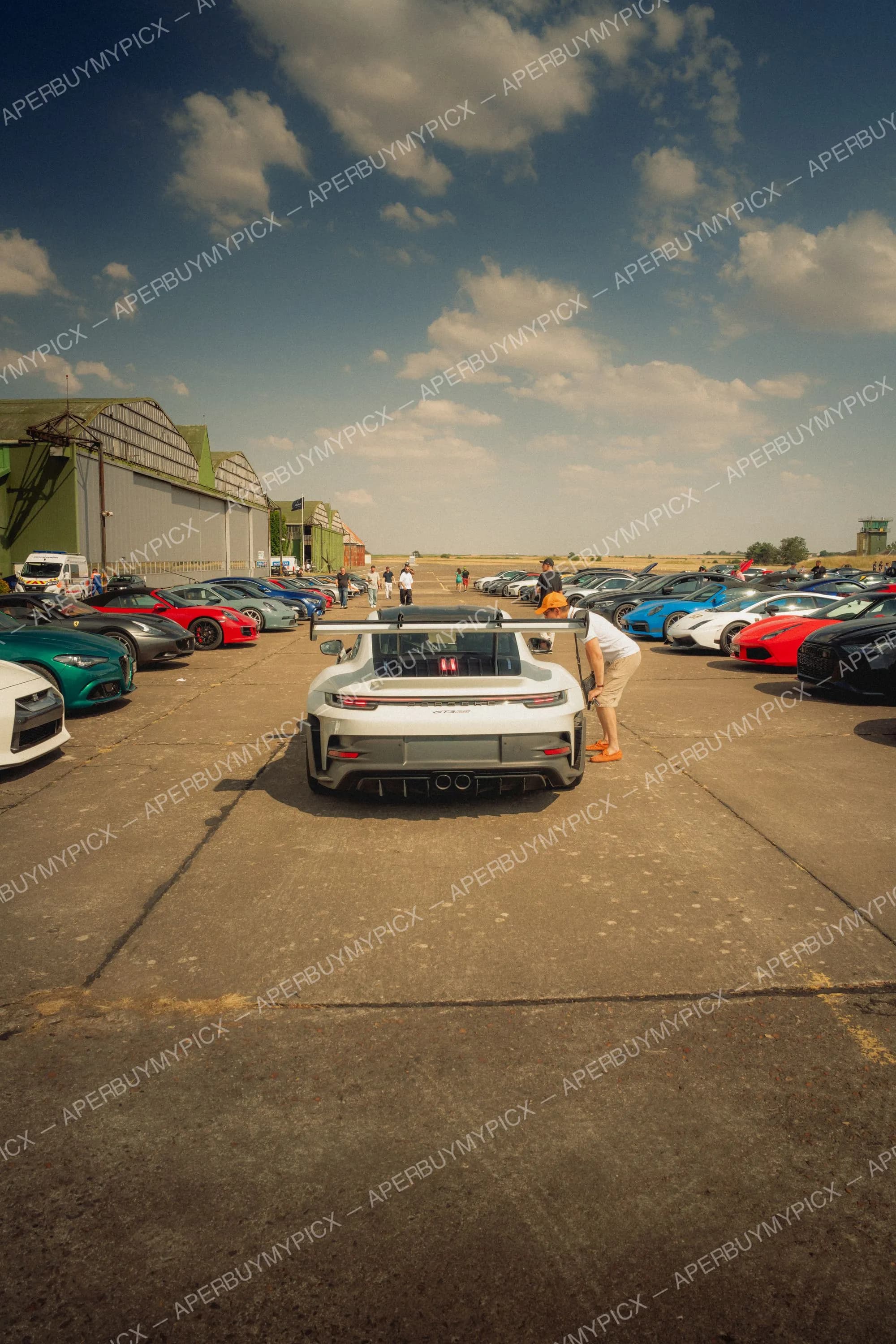 Porsche 911 GT3 RS grise vue de l'arrière avec aileron, garée sur une piste d'aérodrome entourée de nombreuses supercars colorées, Ferrari, Lamborghini et Alfa Romeo, sous un ciel bleu nuageux avec hangars en arrière-plan.