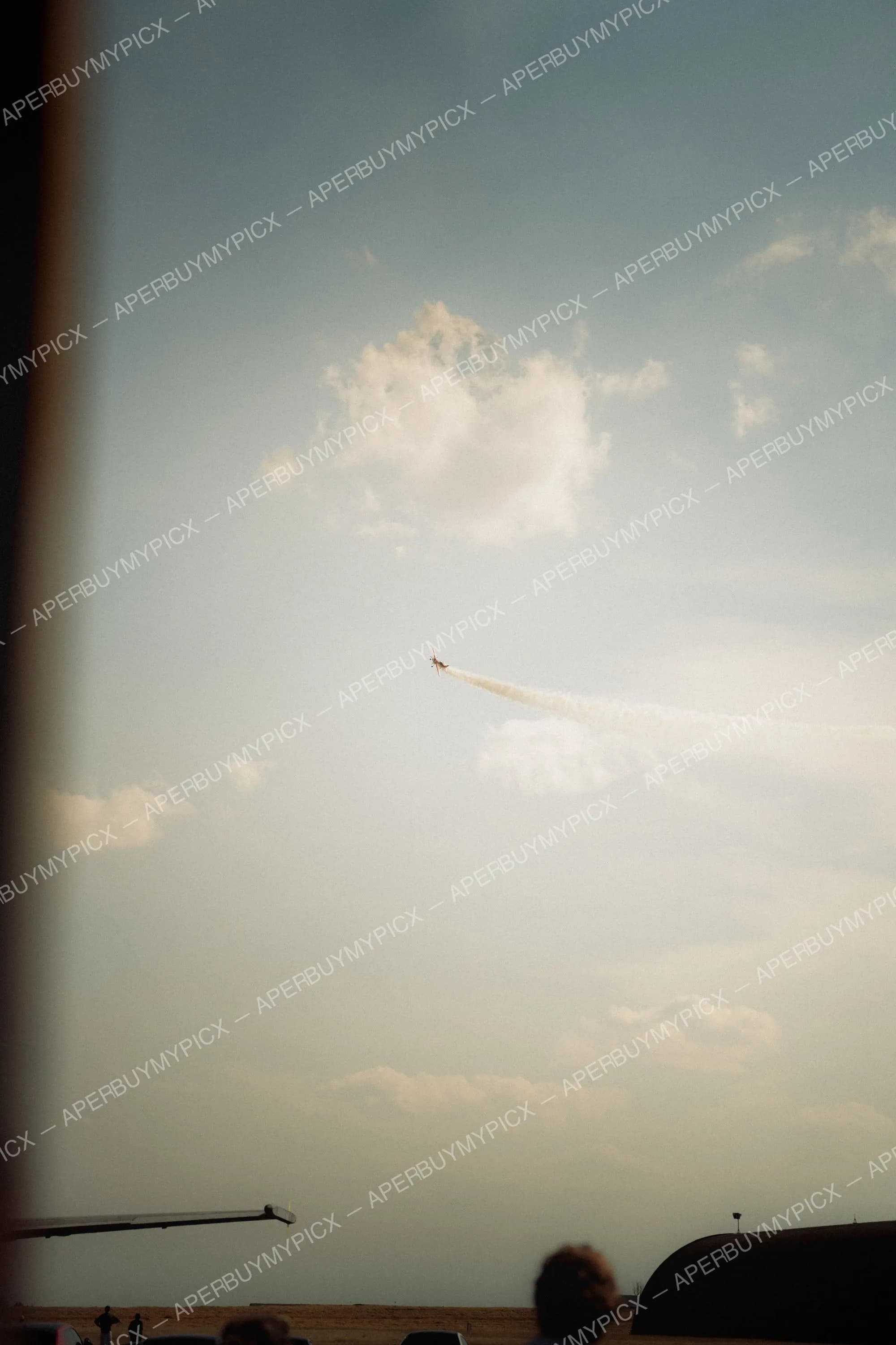 Avion de voltige laissant une traînée de fumée blanche dans un ciel bleu partiellement nuageux lors d'un meeting aérien, spectateurs au sol.