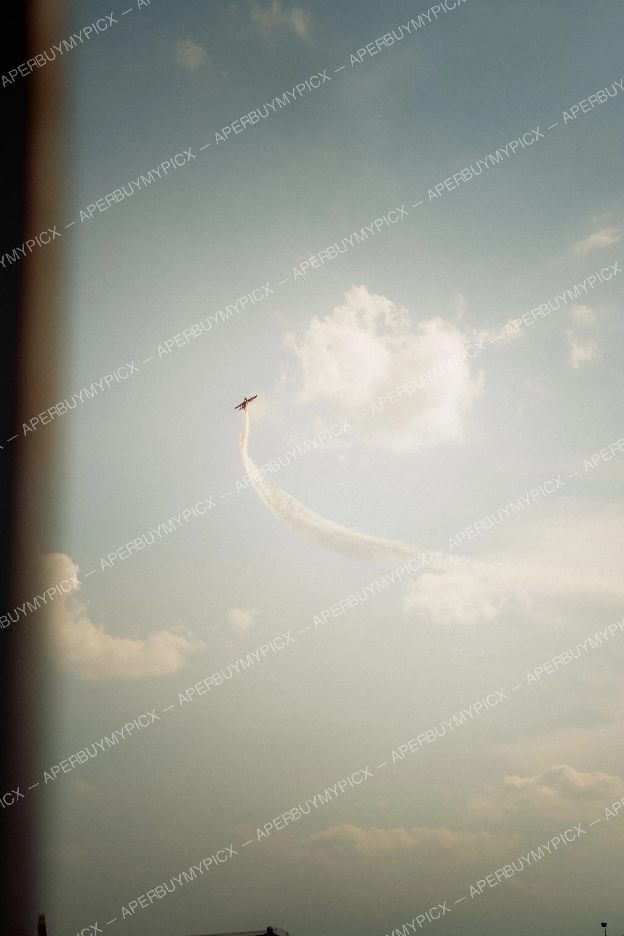 Petit avion de voltige rouge et blanc effectuant une figure acrobatique, laissant une traînée de fumée courbée dans un ciel bleu parsemé de nuages blancs.