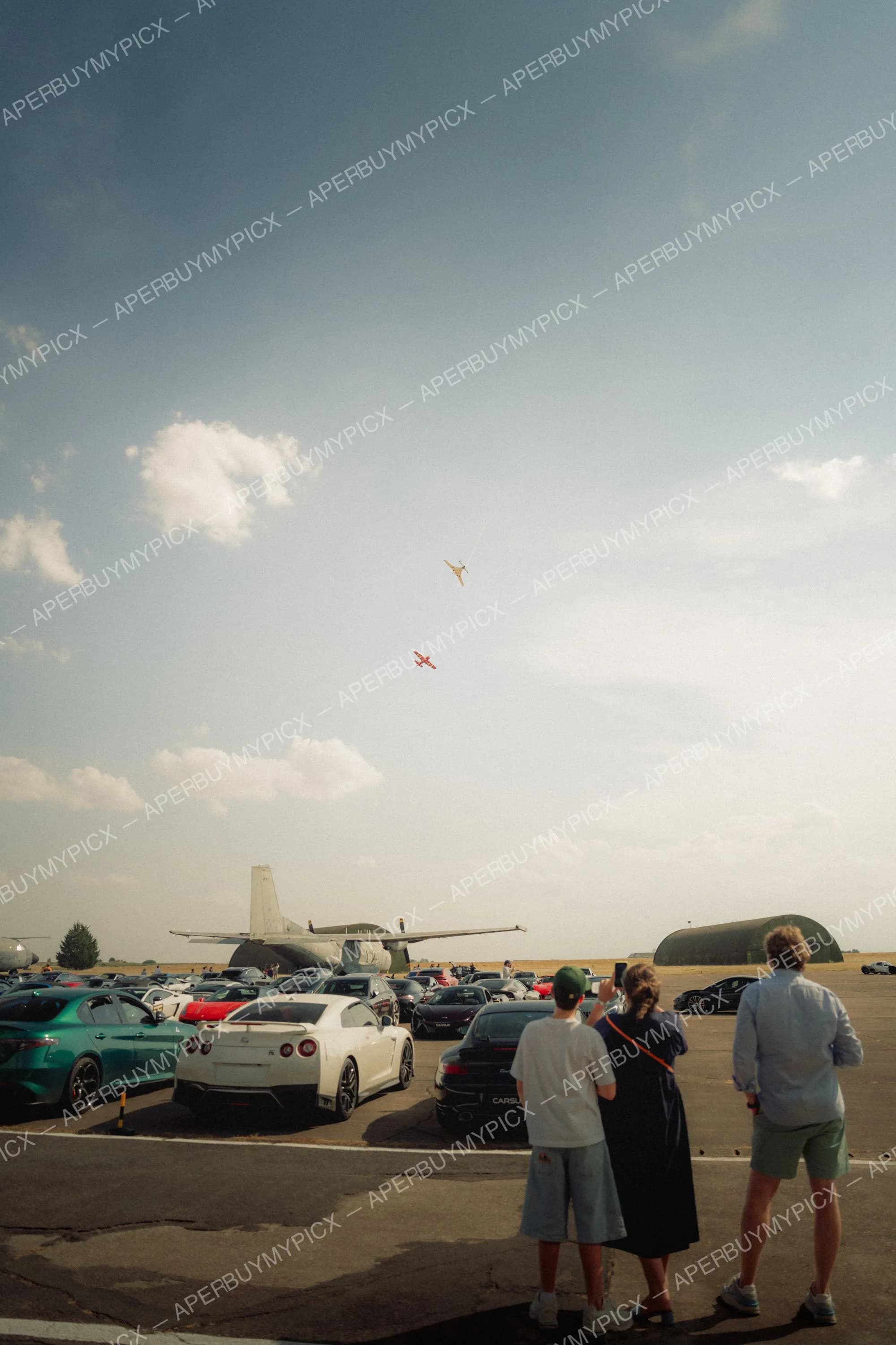 Spectateurs observant deux avions en vol au-dessus d'un tarmac rempli de voitures de sport et d'un avion militaire, sous un ciel nuageux.
