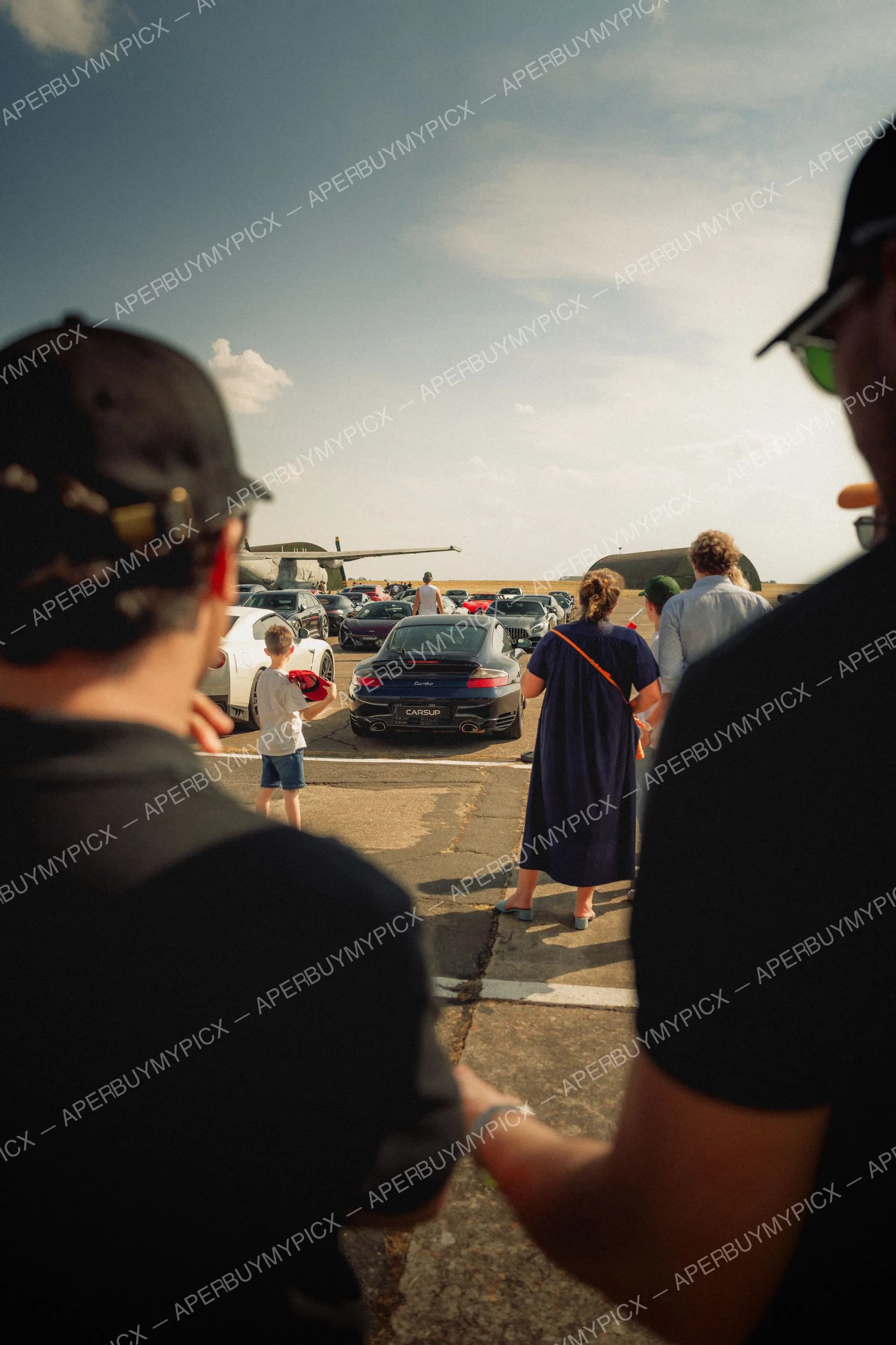 Spectateurs observant une Porsche 911 Turbo noire et d'autres voitures de sport garées sur une piste d'aéroport, avec un avion militaire et des hangars en arrière-plan sous un ciel ensoleillé.