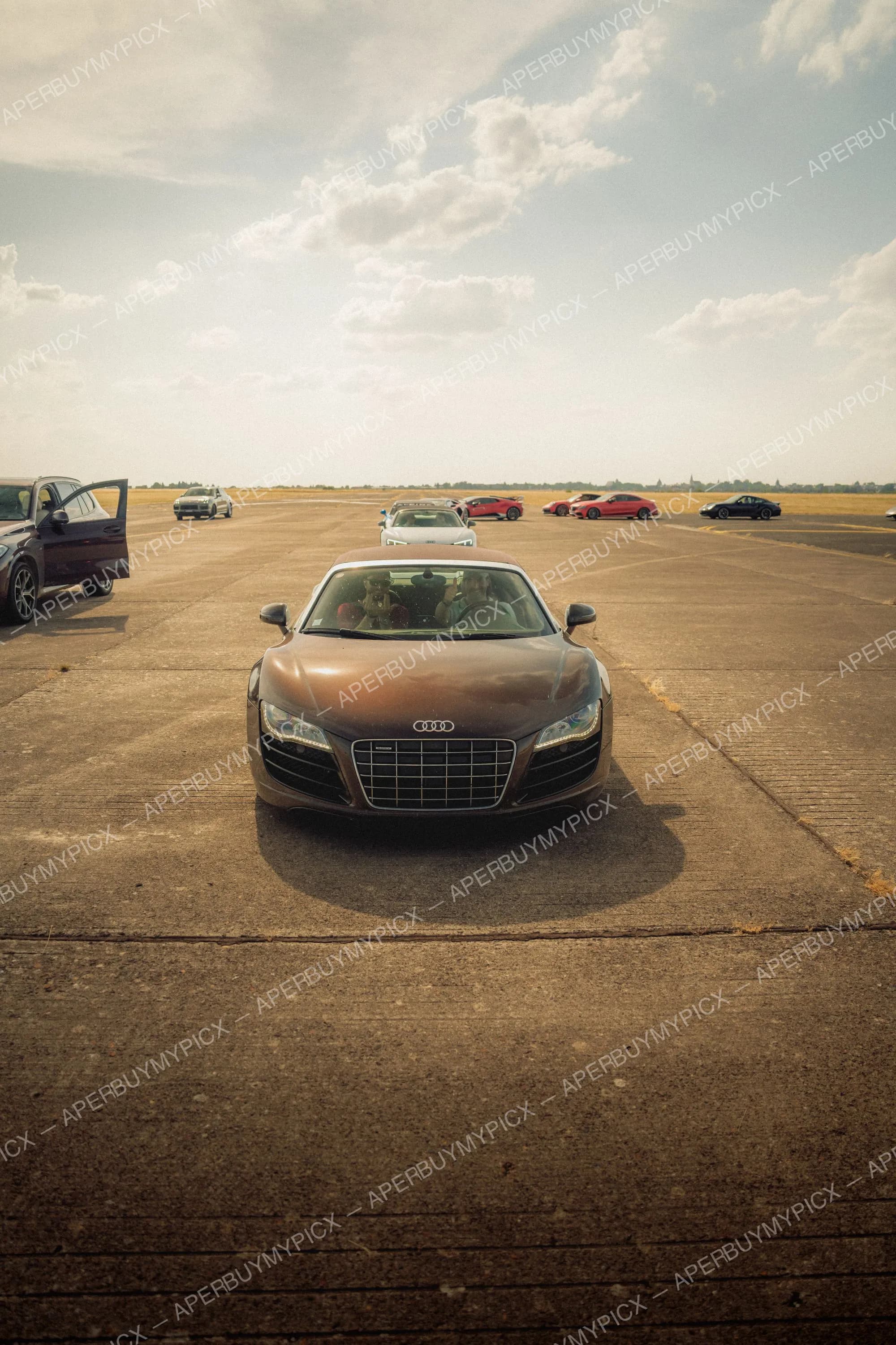 Audi R8 Spyder brune vue de face garée sur une piste d'aérodrome ensoleillée, avec plusieurs voitures de sport rouges et noires en arrière-plan sous un ciel nuageux.