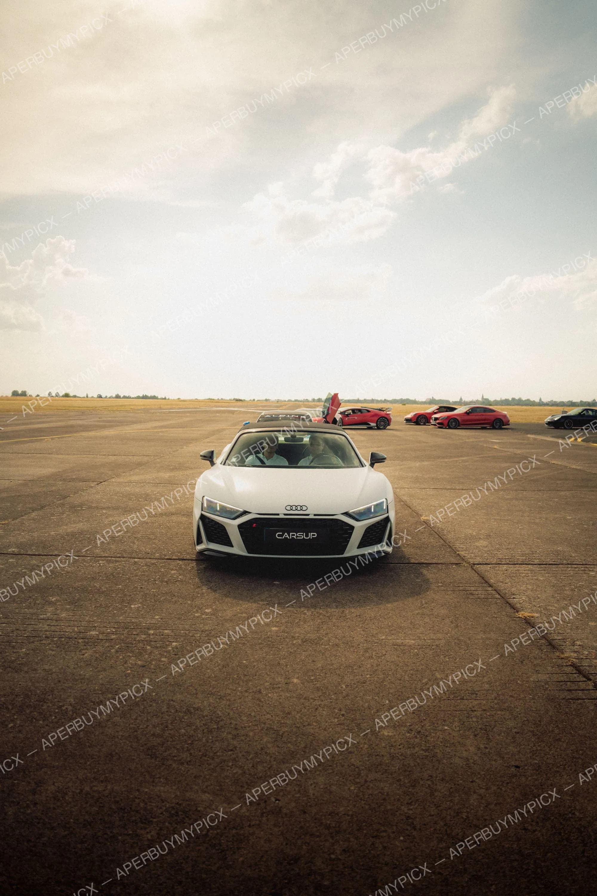 Audi R8 Spyder blanche décapotable roulant sur une piste d'aérodrome, avec deux occupants à bord, sous un ciel nuageux et des supercars rouges garées en arrière-plan.
