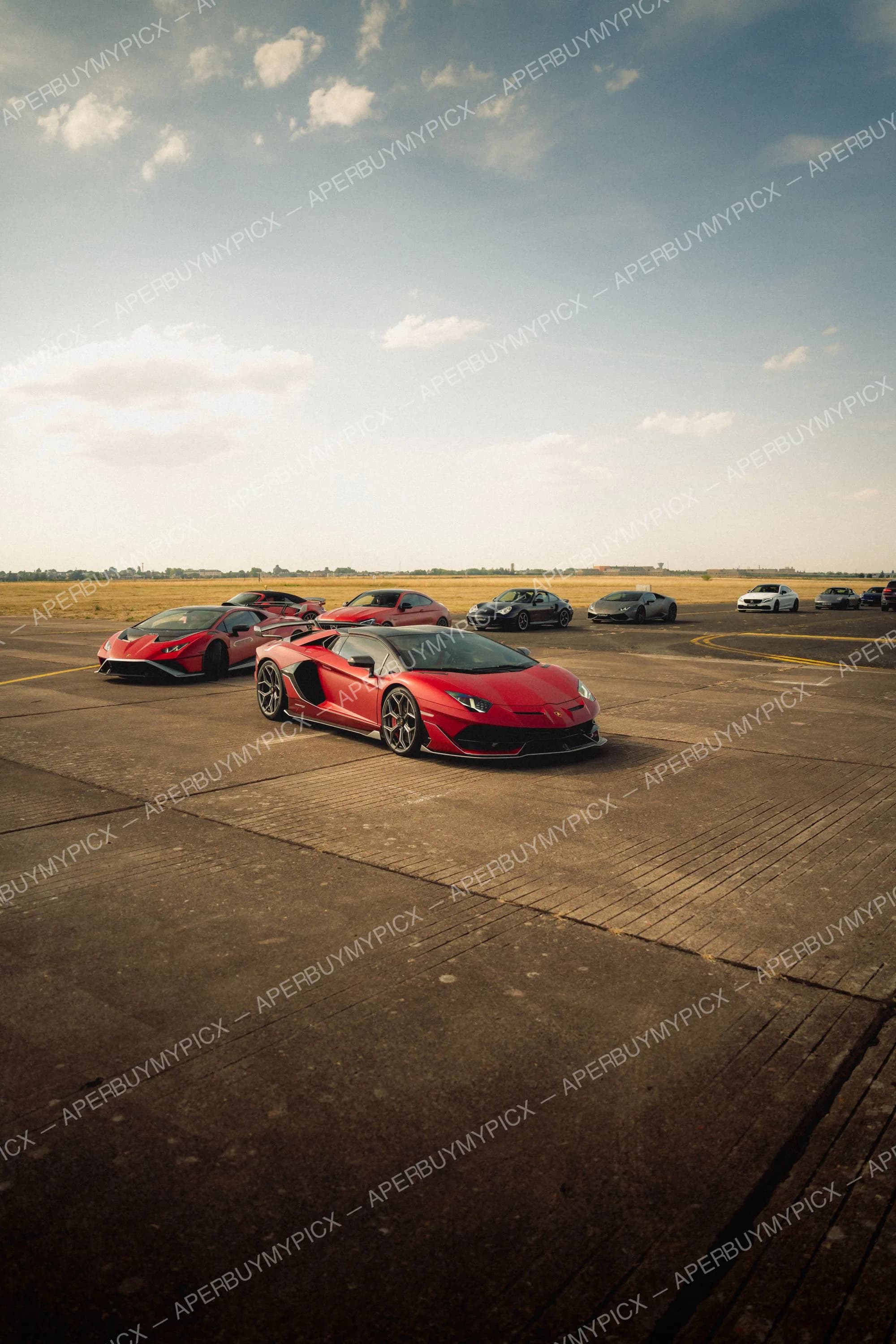 Lamborghini Aventador SVJ rouge au premier plan sur une piste d'aéroport, entourée de Huracan rouges, Porsche 911, Mercedes et autres supercars sous un ciel nuageux.