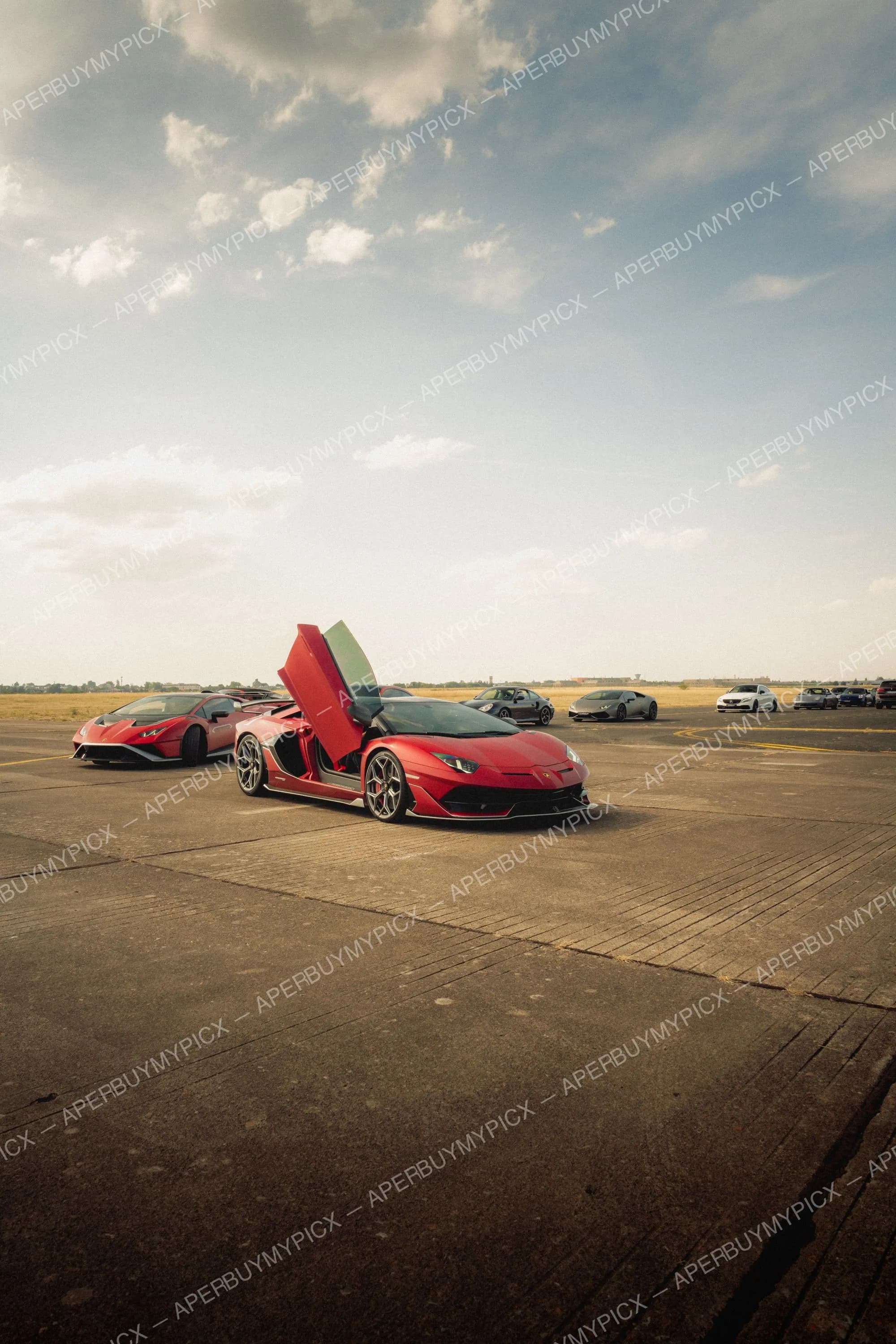 Lamborghini Aventador SVJ rouge avec porte papillon ouverte sur le tarmac d'un ancien aéroport, entourée d'une Huracán rouge, d'une Huracán grise, d'une Porsche 911 et d'autres voitures sous un ciel nuageux.