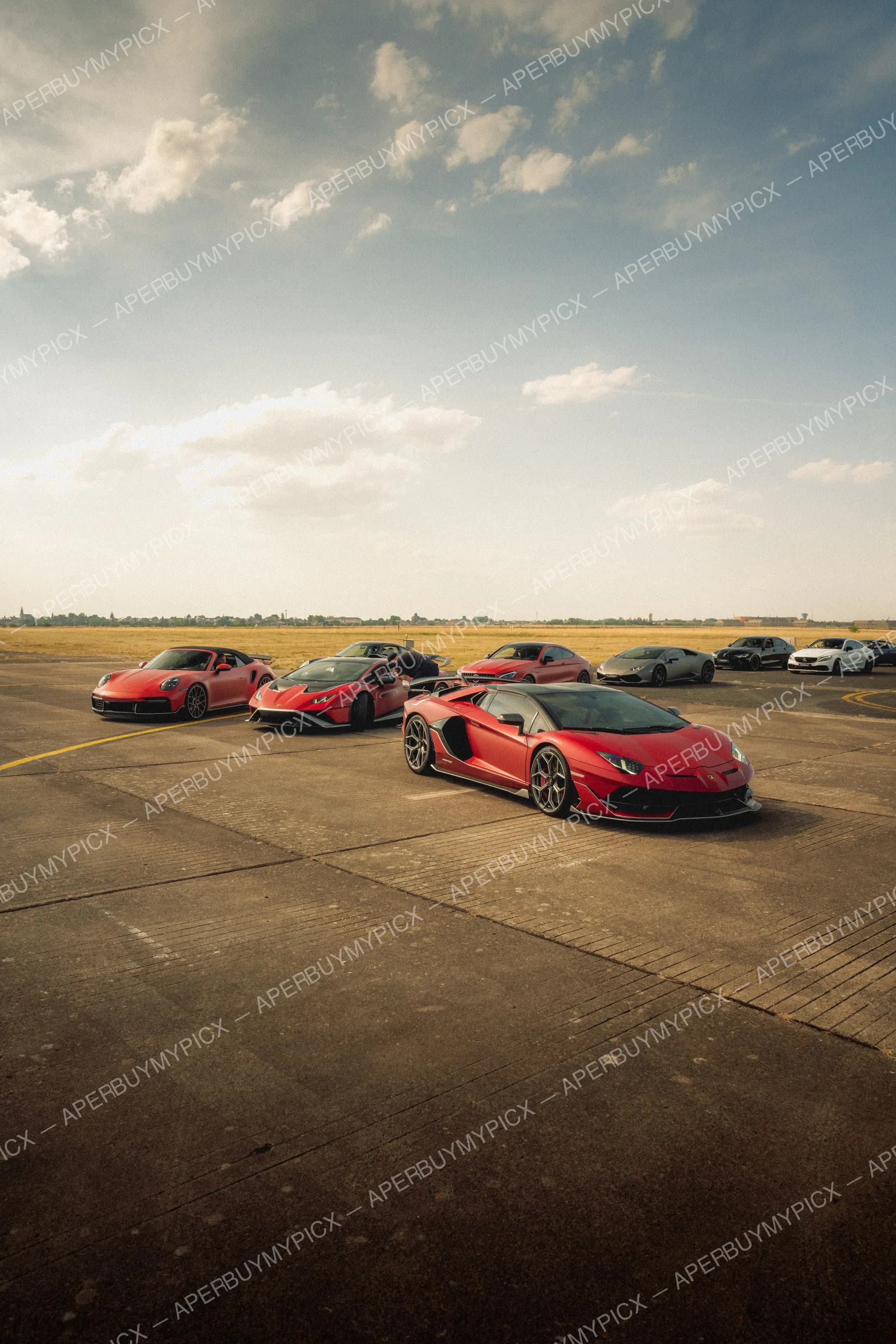 Lamborghini Aventador SVJ rouge au premier plan avec Huracán, Porsche 911 et autres voitures de sport alignées sur le tarmac d'un aéroport sous un ciel nuageux