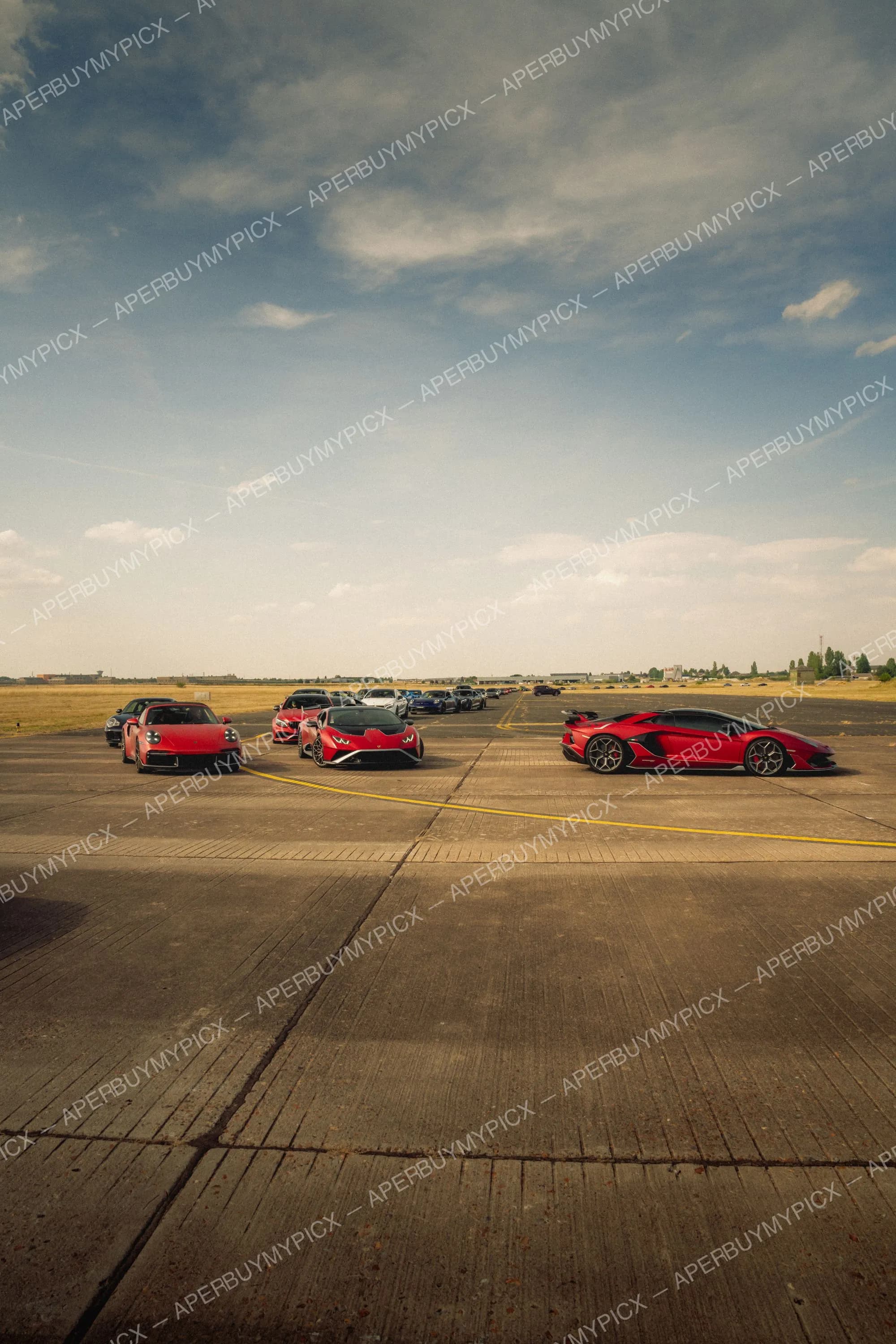 Trois supercars rouges dont une Porsche 911, une Lamborghini Huracan et une Aventador SVJ garées sur une piste d'aéroport avec un ciel nuageux et d'autres voitures en arrière-plan