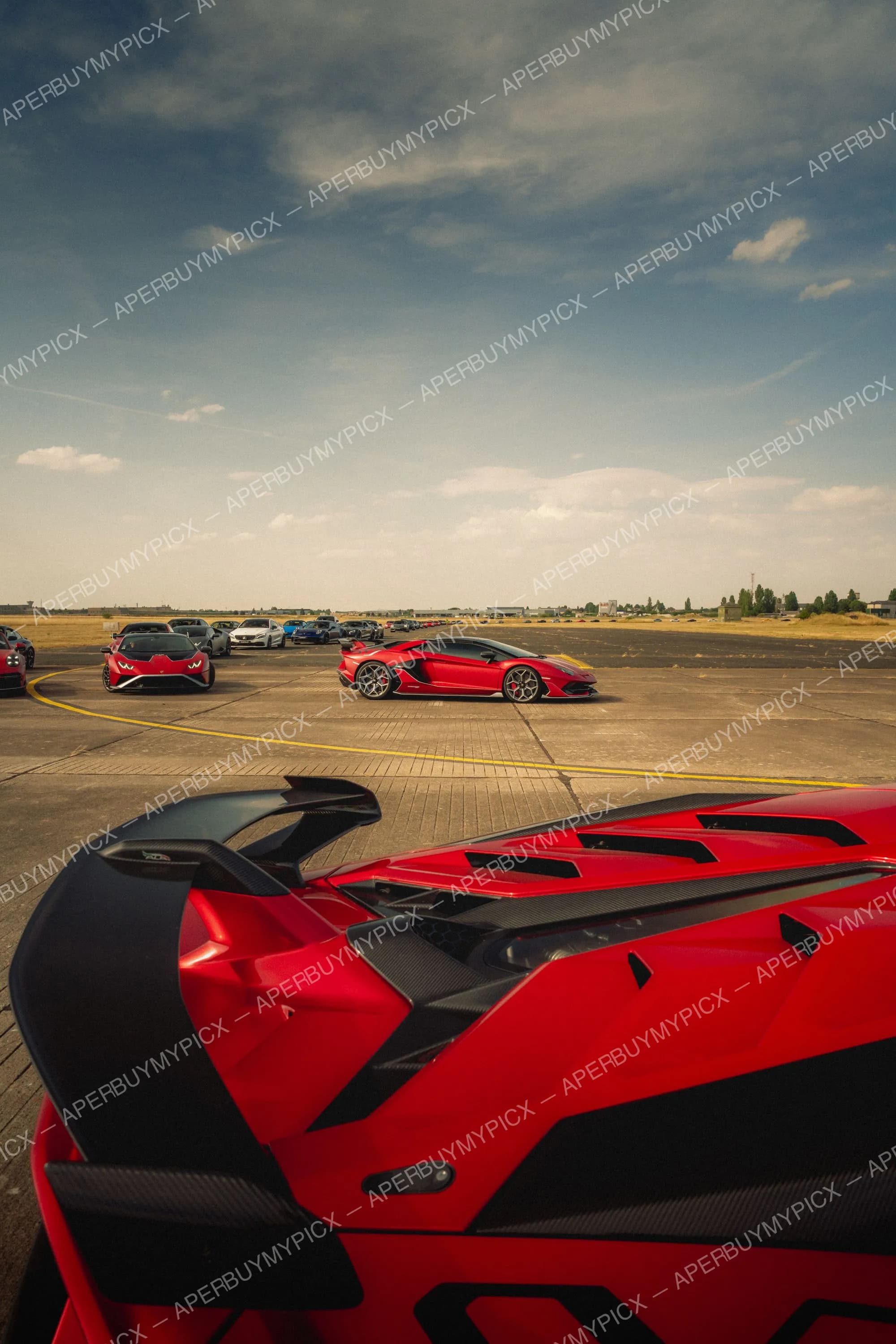 Lamborghini Aventador SVJ rouge avec aileron carbone au premier plan, une seconde SVJ rouge et une Huracán rouge sur une piste d'aéroport sous un ciel nuageux