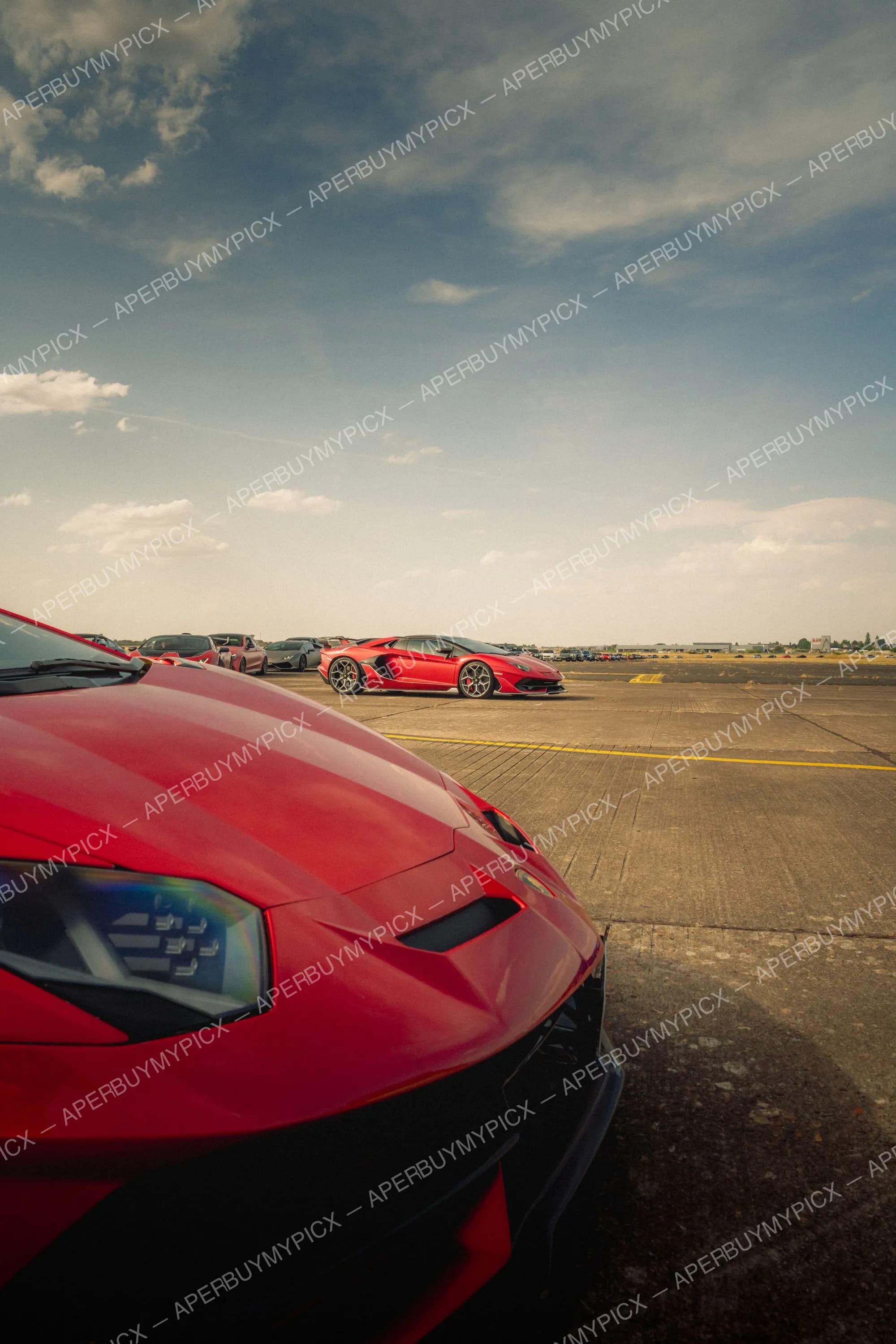 Deux Lamborghini Aventador SVJ rouges garées sur une piste d'aéroport sous un ciel nuageux, avec d'autres supercars alignées à l'arrière-plan