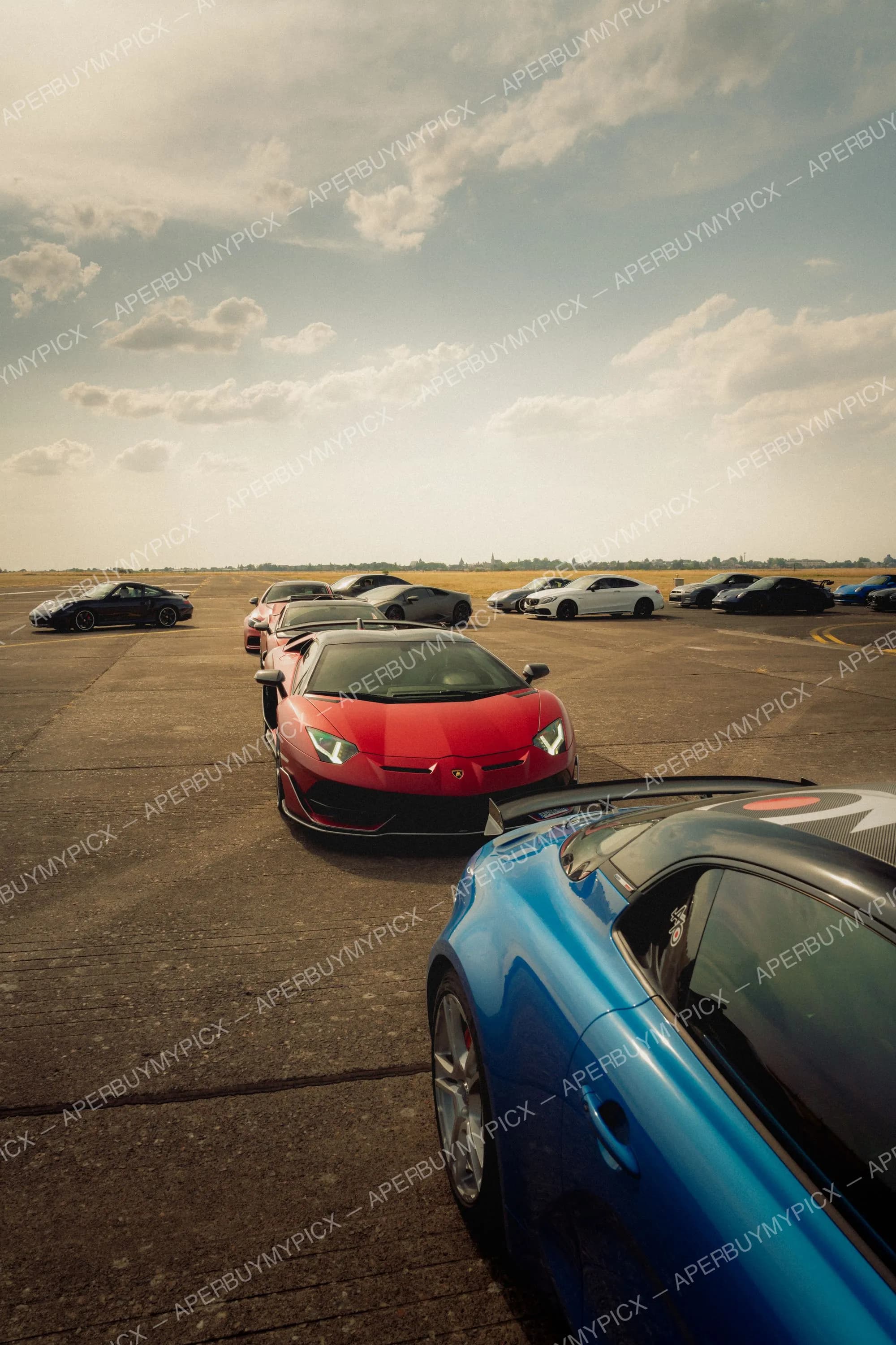 Rassemblement de supercars sur une piste d'aérodrome au coucher du soleil : Lamborghini Aventador SVJ rouge au centre, Porsche bleue au premier plan, Porsche 911, Mercedes AMG blanche et Nissan GT-R alignées à l'horizon sous un ciel nuageux.