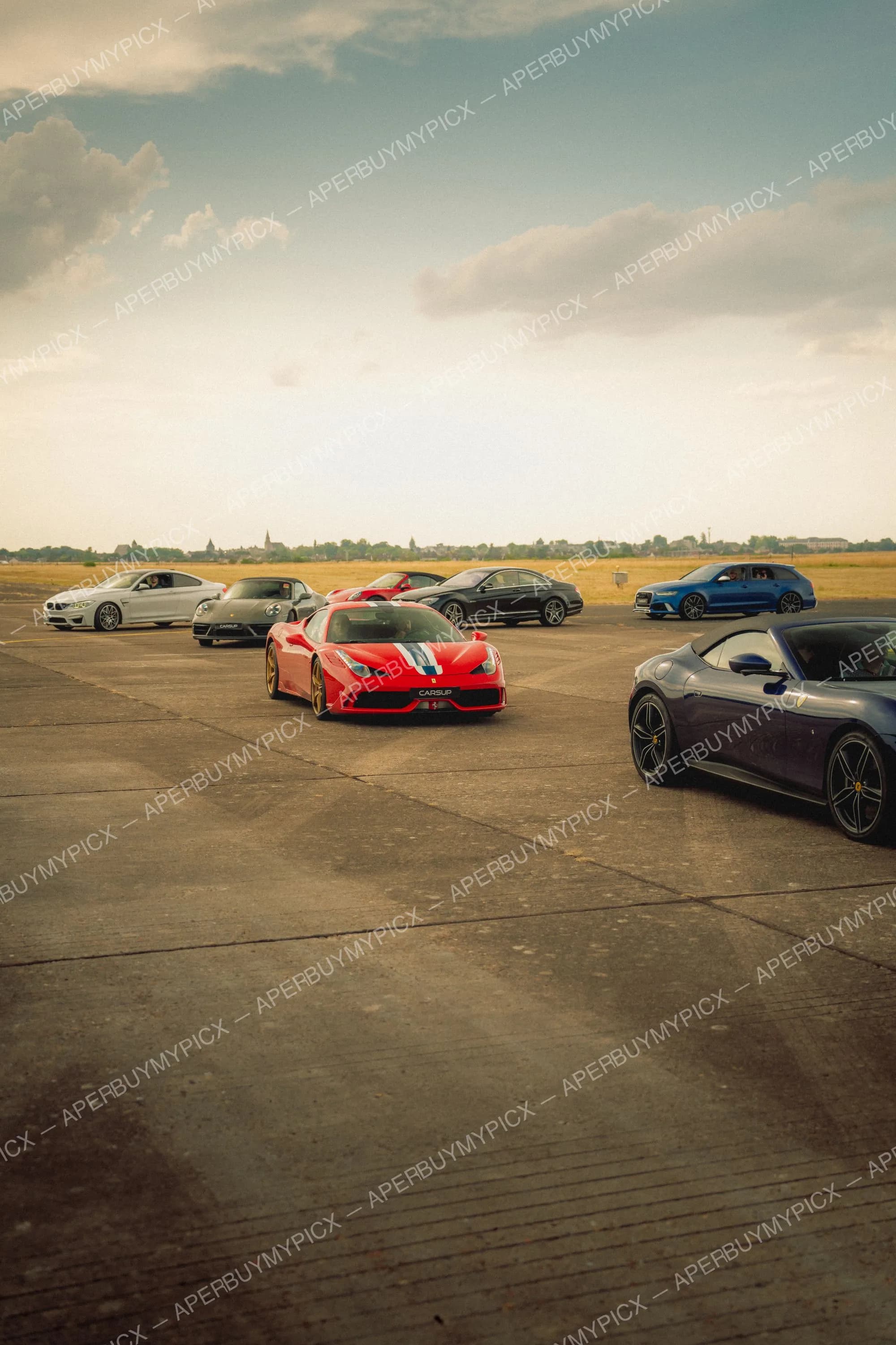 Ferrari 458 Speciale rouge à bande bleue au centre d'un rassemblement de supercars sur une piste d'aérodrome au coucher du soleil, avec une Ferrari cabriolet bleue, une BMW blanche, une Porsche noire, une Audi RS6 bleue et une Mercedes noire.