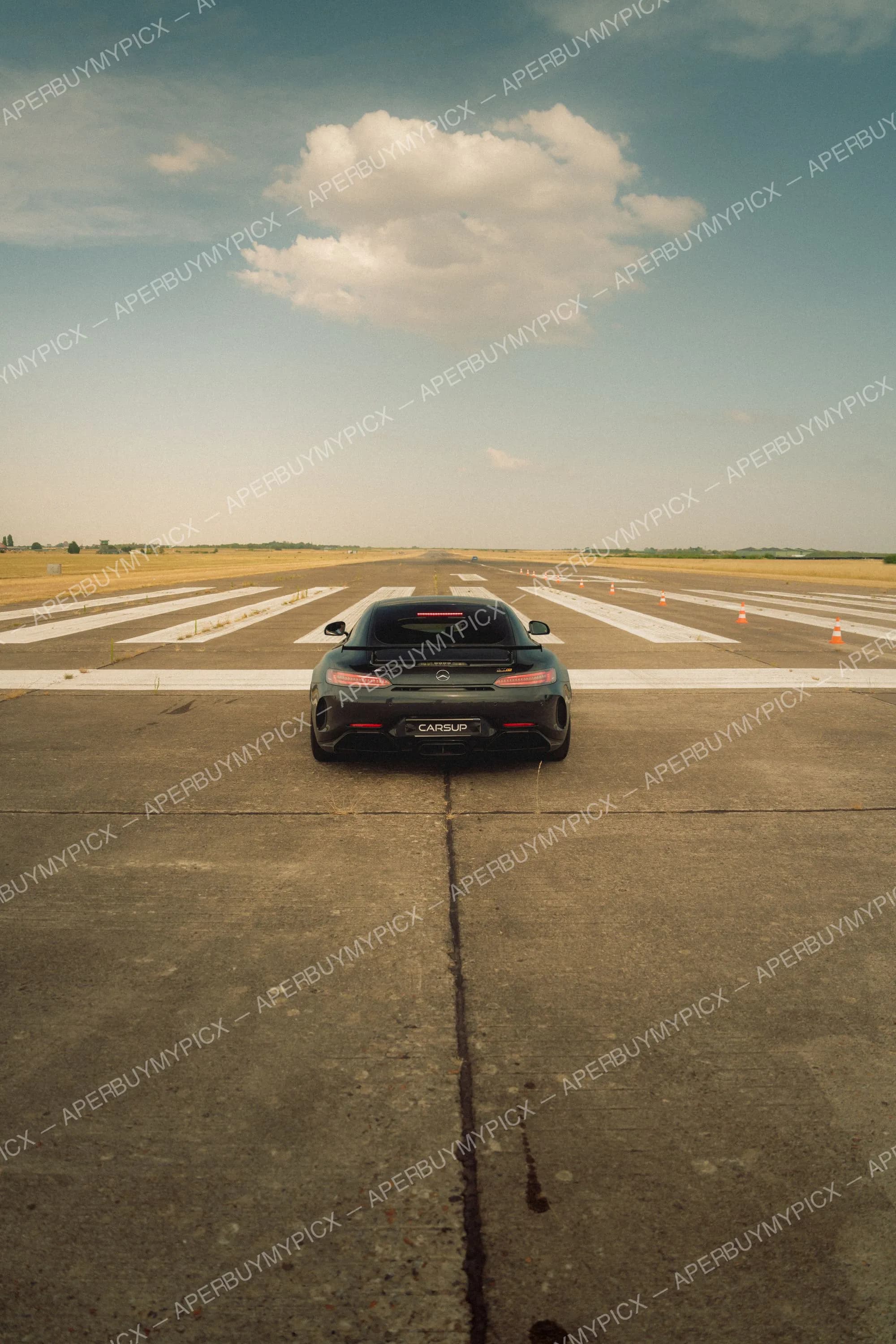 Vue arrière d'une Mercedes-AMG GT R noire garée au bout d'une piste d'aéroport bordée de cônes orange, sous un ciel bleu partiellement nuageux.