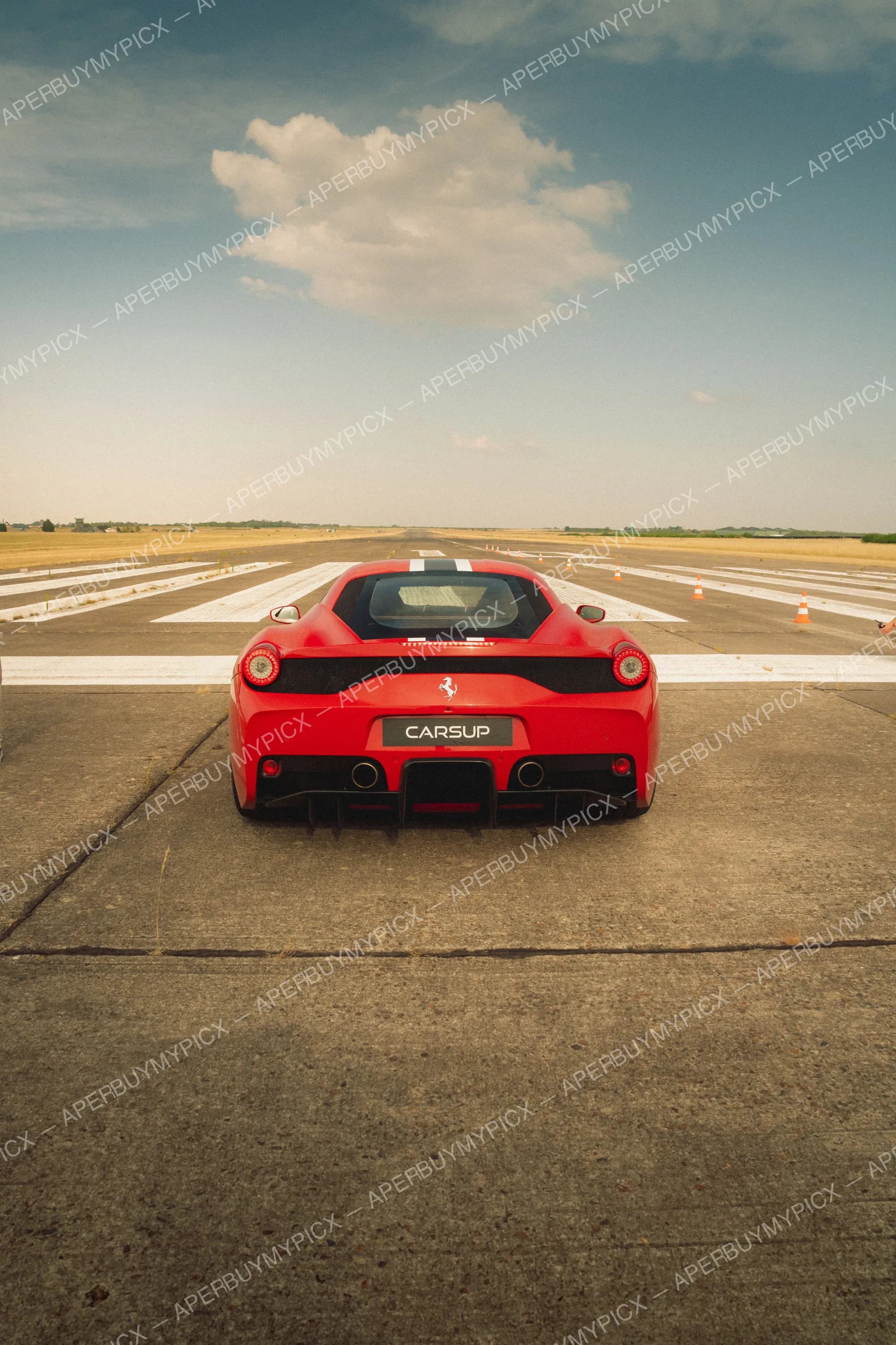 Vue arrière d'une Ferrari 458 Speciale rouge avec bandes blanches, garée sur une piste d'aéroport avec des cônes orange sous un ciel bleu nuageux.