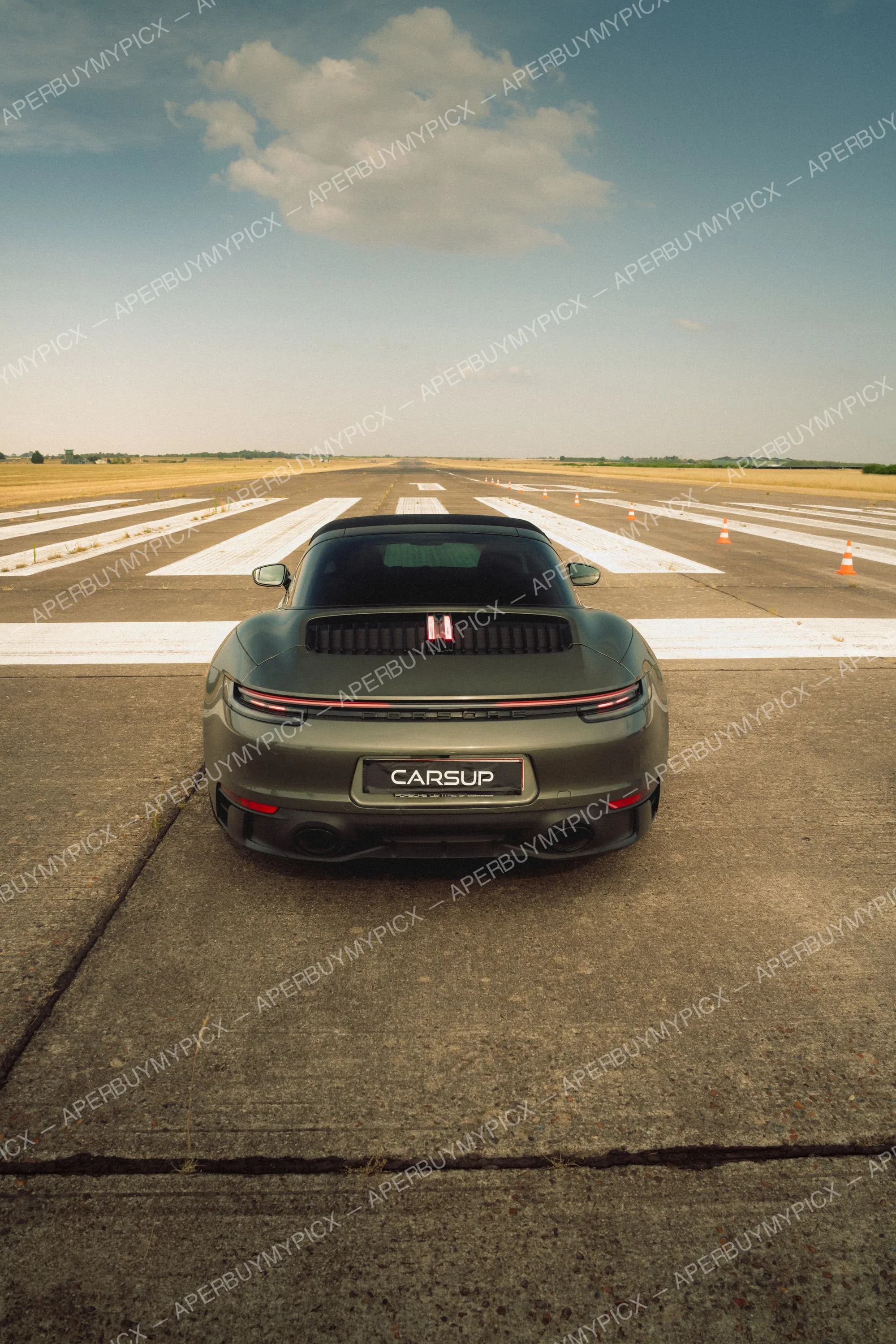 Vue arrière d'une Porsche 911 vert kaki stationnée au centre d'une piste d'aviation, avec cônes orange et ciel nuageux en arrière-plan