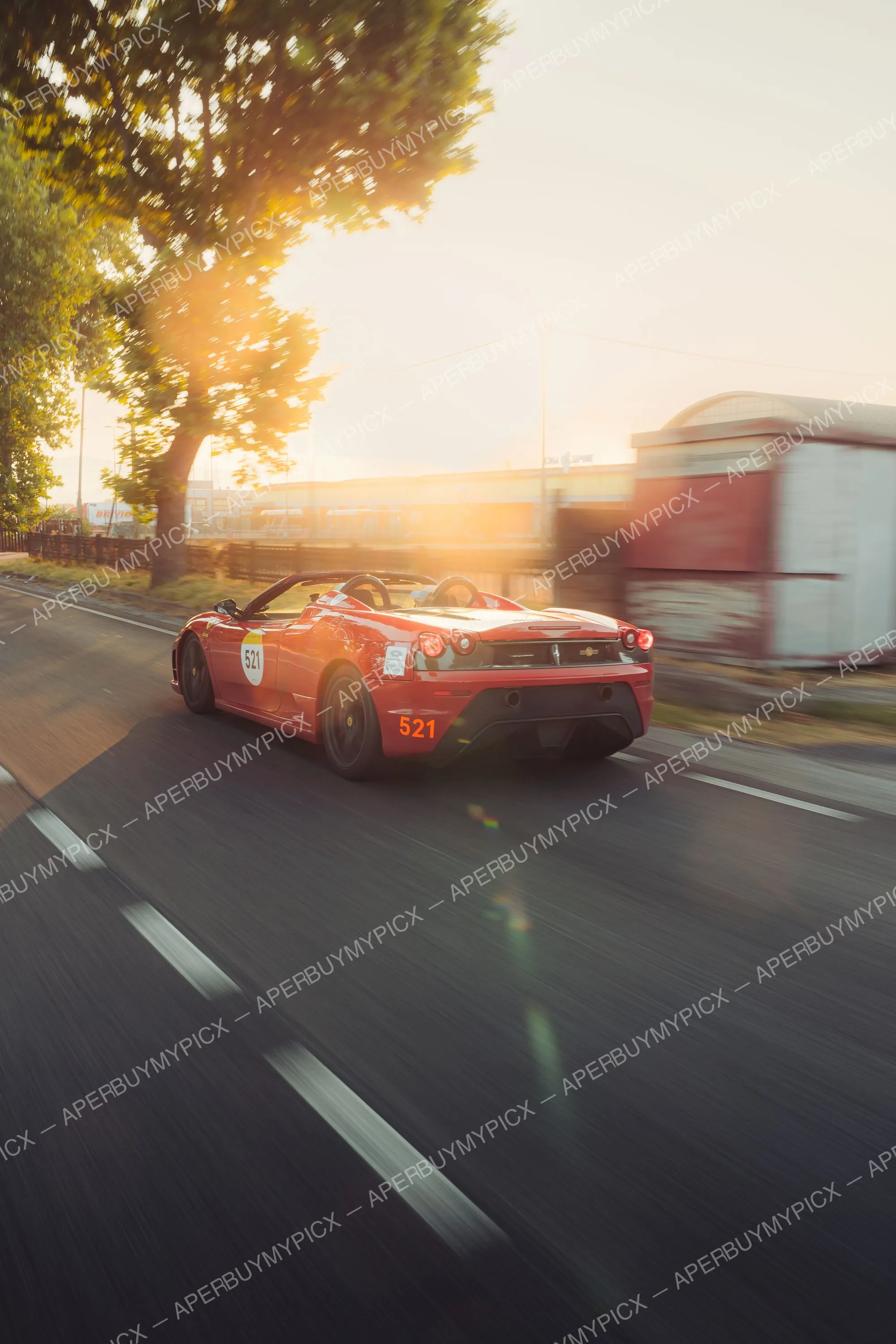 Ferrari 430 Scuderia Spider rouge numéro 521 roulant à vive allure sur une route au coucher de soleil, vue arrière avec effet de mouvement, arbres et bâtiments industriels en arrière-plan.