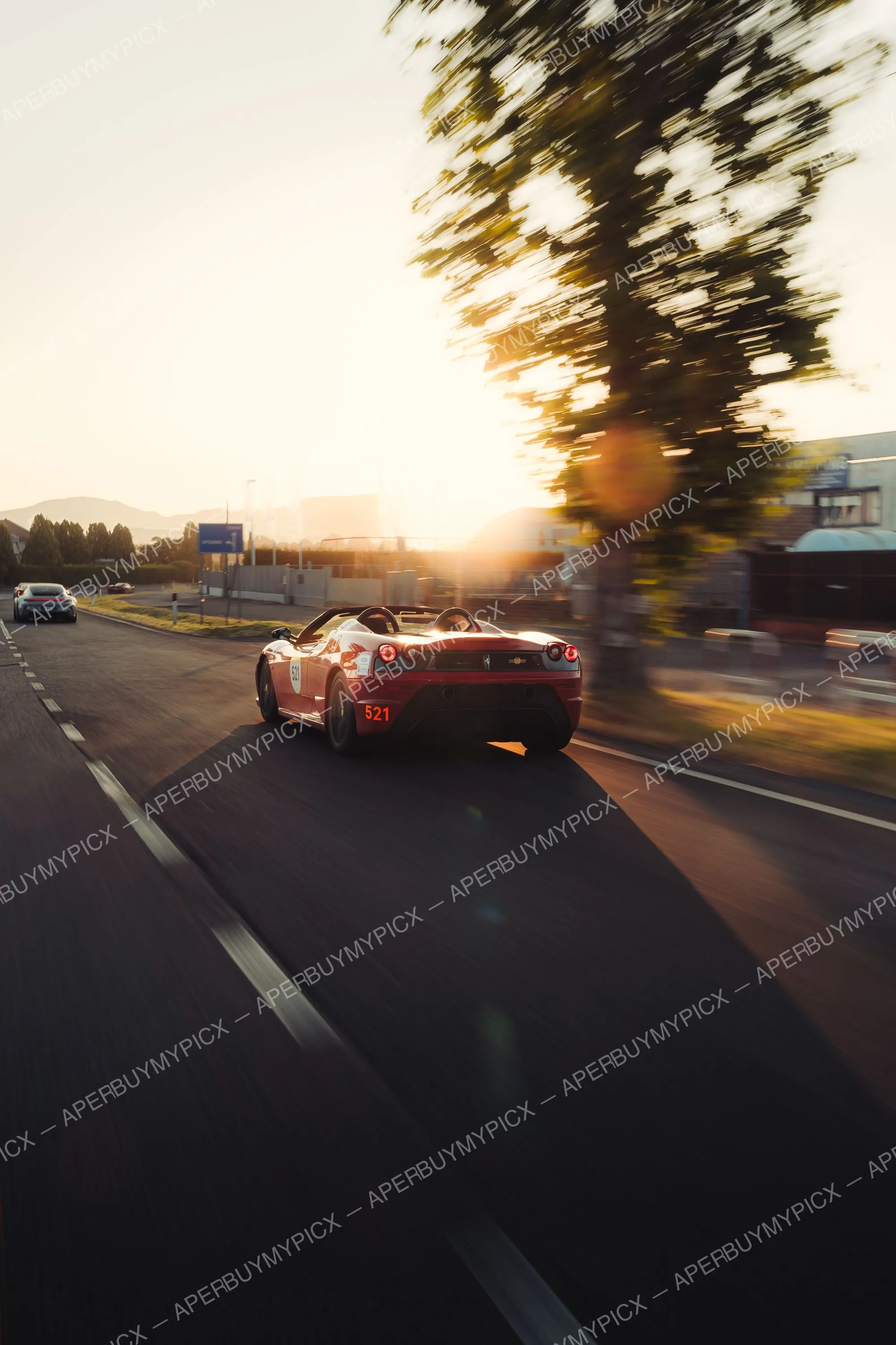 Ferrari 430 Scuderia Spider rouge numéro 521 roulant sur une route au coucher du soleil, vue arrière, avec effet de vitesse et flare solaire, lors de la Mille Miglia 2025.