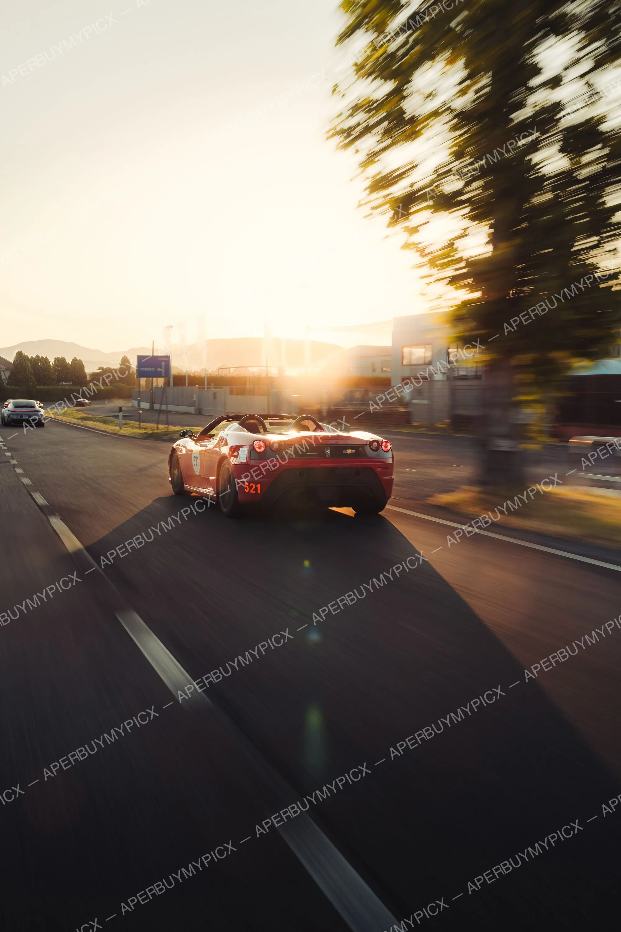 Ferrari Spider rouge et blanche numéro 521 roulant sur une route au coucher du soleil lors de la Mille Miglia 2025, avec une Porsche au loin et un arbre flou en mouvement à droite.