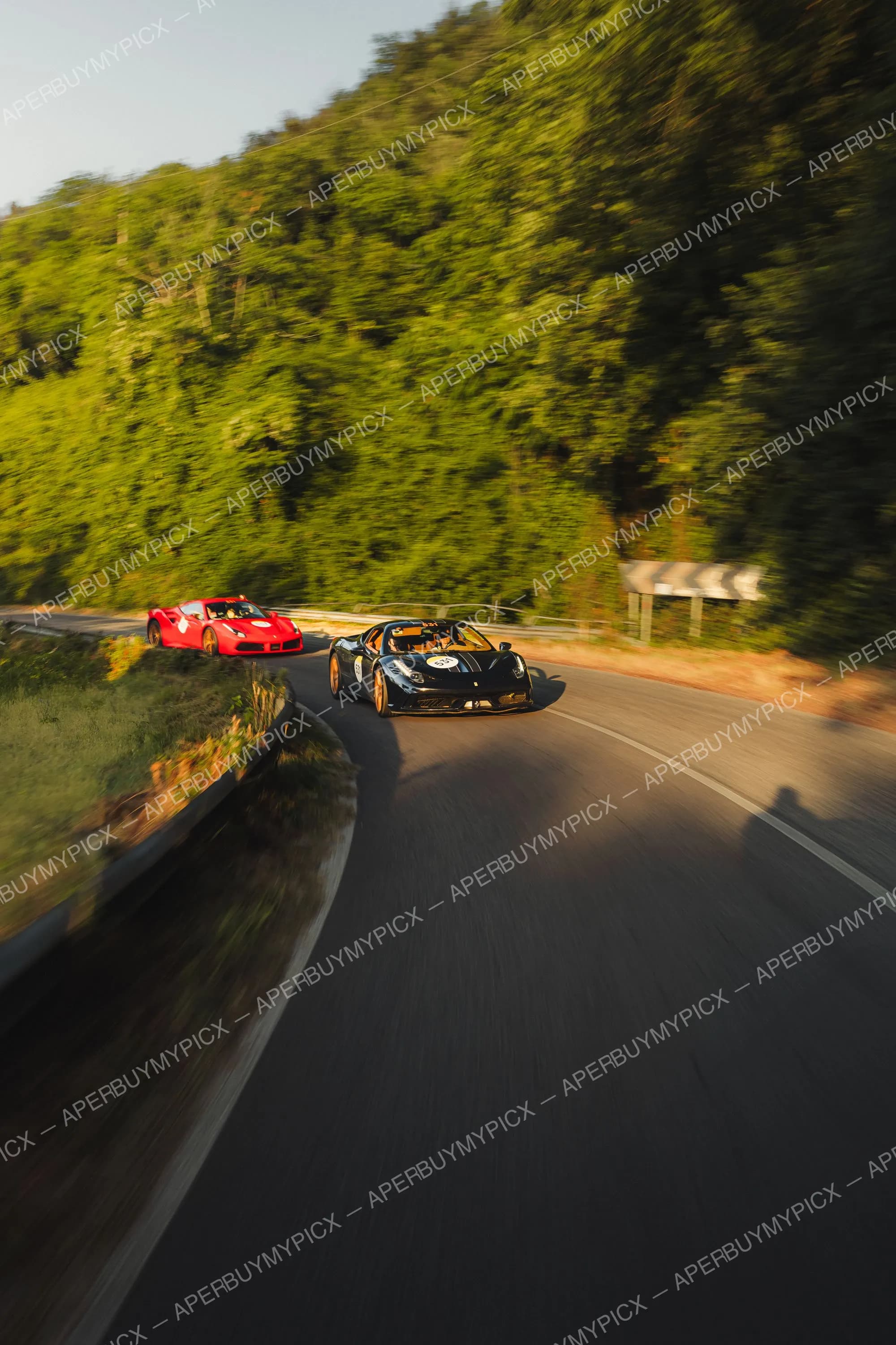 Une Ferrari noire numéro 531 et une Ferrari rouge roulant à vive allure sur une route de montagne sinueuse bordée d'une forêt verdoyante, effet de filé au coucher du soleil, Mille Miglia 2025.