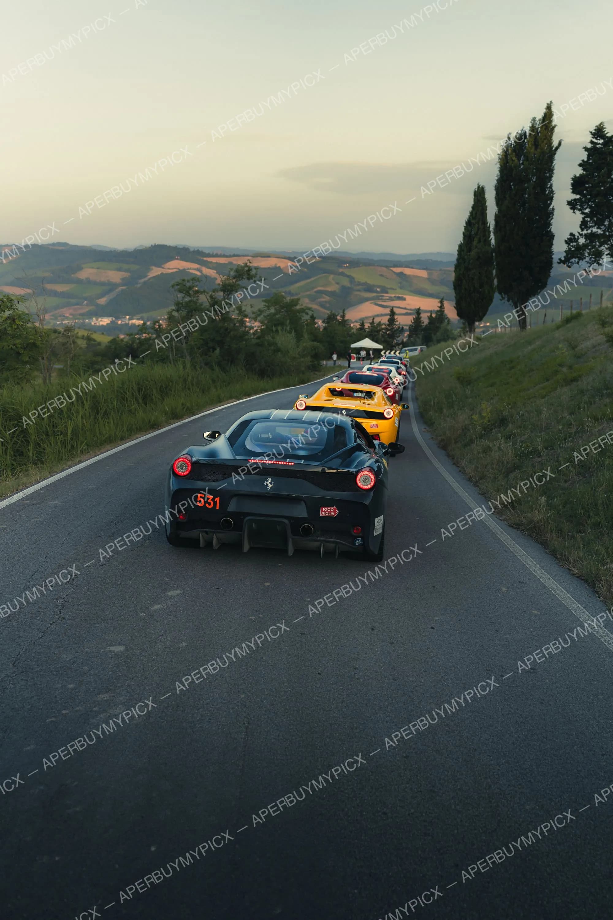 Ferrari 458 Speciale noire numéro 531 vue de l'arrière suivie de Ferrari jaune et rouge sur une route sinueuse au coucher du soleil dans les collines de Toscane lors de la Mille Miglia 2025