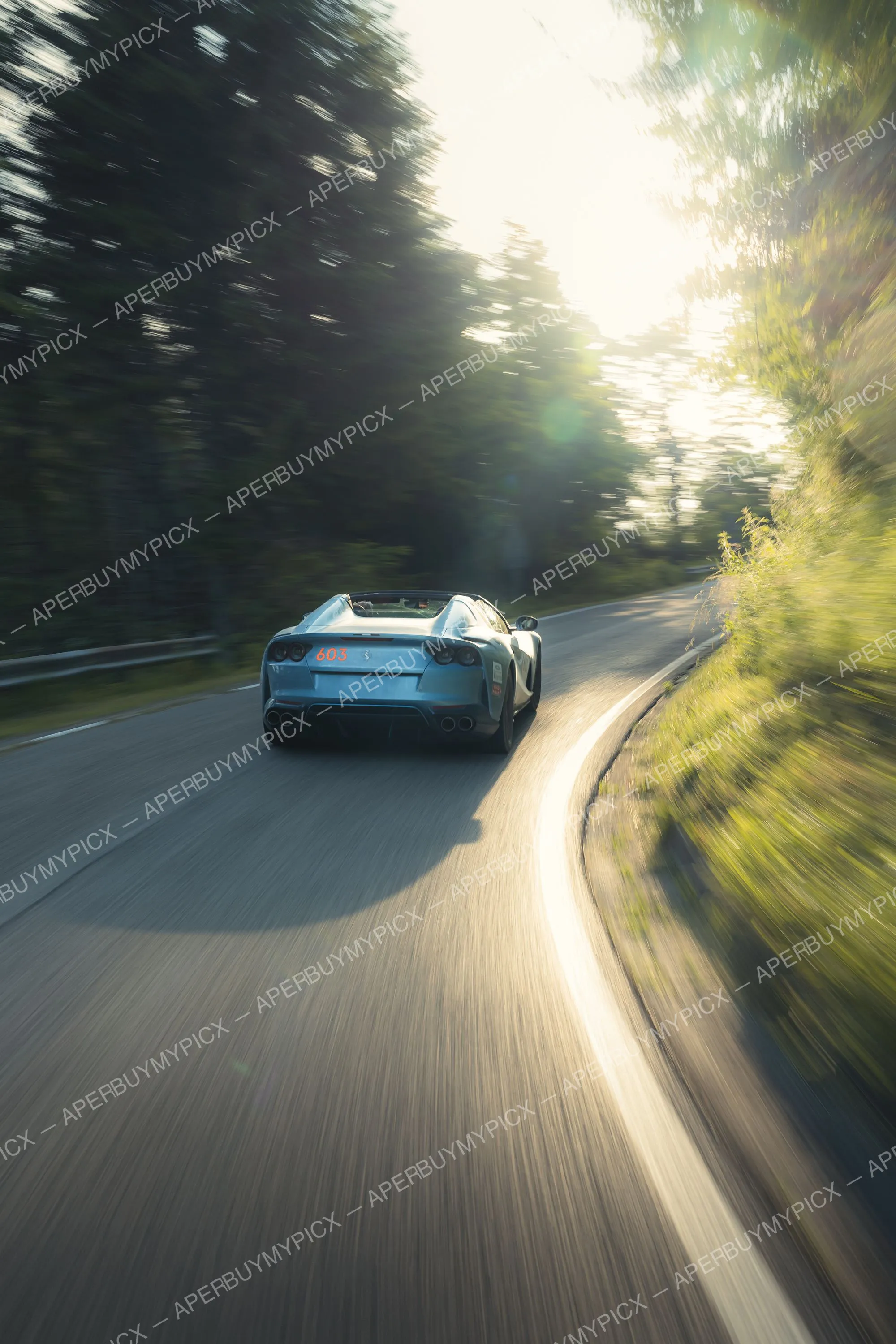 Ferrari 812 GTS bleue numéro 603 vue de l'arrière filant à grande vitesse sur une route sinueuse bordée d'arbres au coucher du soleil, effet de filé