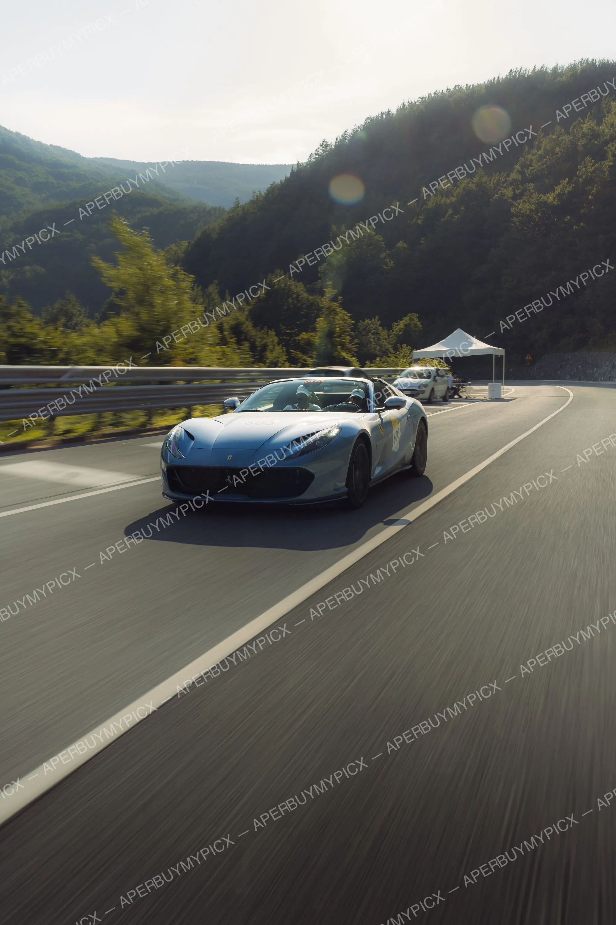 Ferrari 812 GTS décapotable bleu ciel roulant à vive allure sur une route de montagne bordée de forêts, effet de vitesse et lumière du soleil, lors de la Mille Miglia 2025.