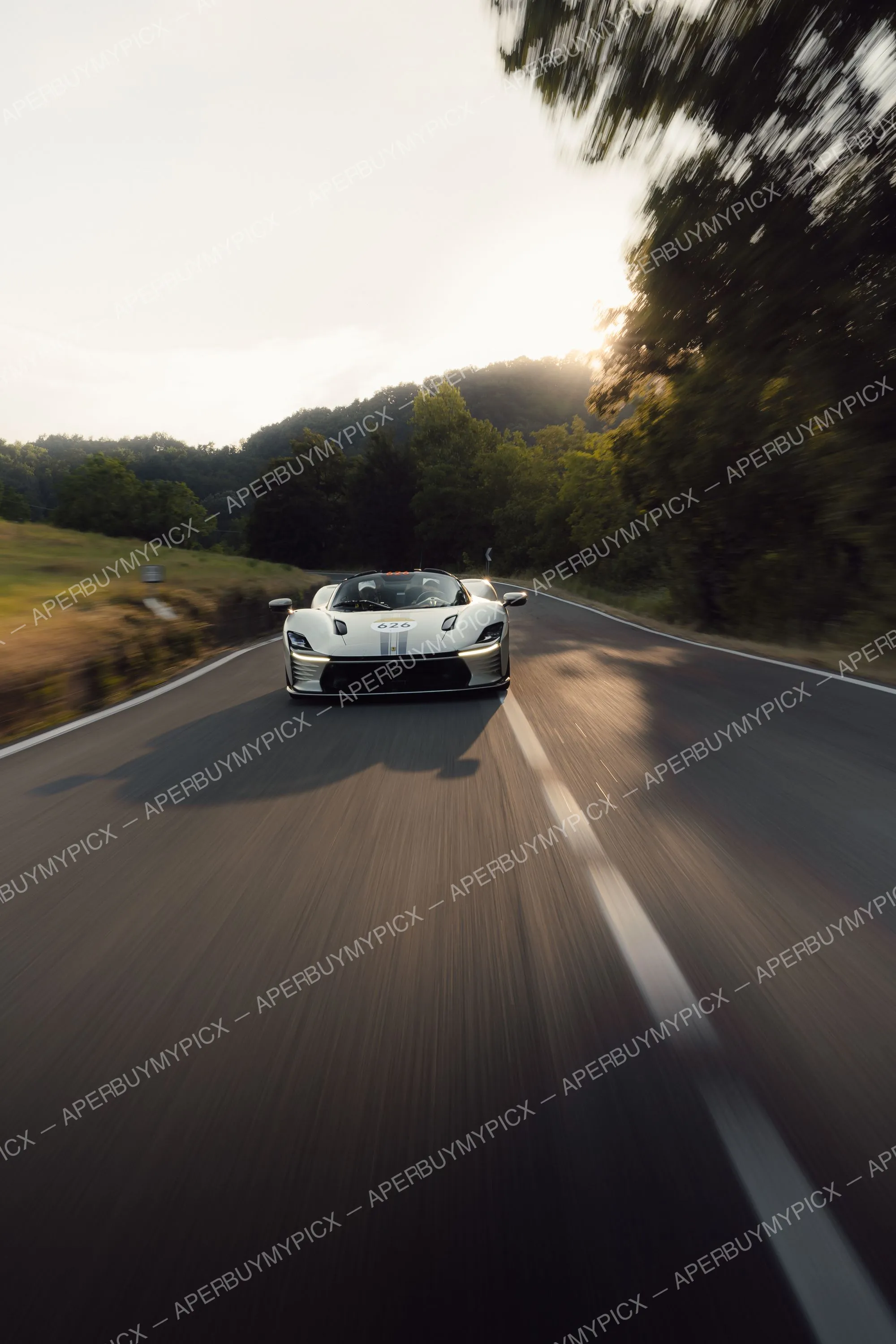 Ferrari Daytona SP3 argentée numéro 626 roulant à vive allure sur une route de campagne bordée d'arbres au coucher du soleil lors des Mille Miglia 2025, effet de flou de vitesse.