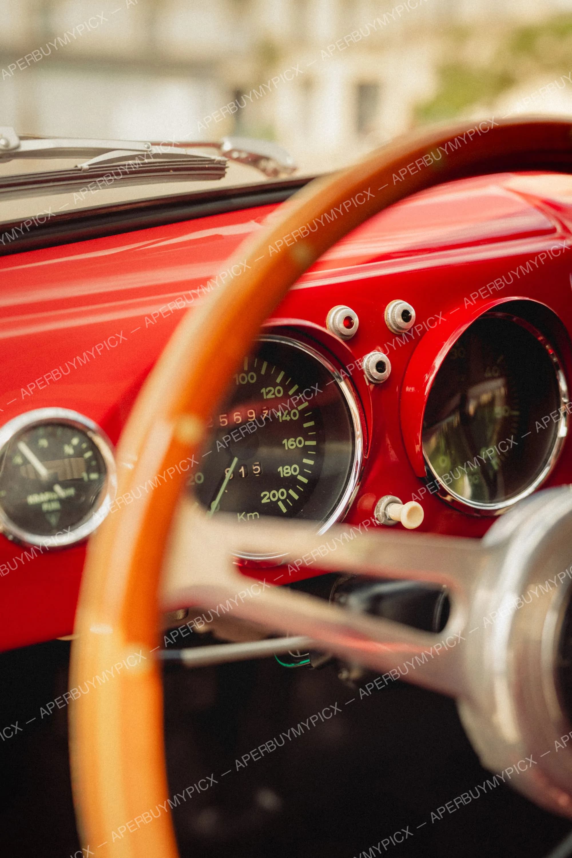 Gros plan sur le tableau de bord rouge d'une Porsche 356 vintage, avec compteur de vitesse gradué jusqu'à 200 km/h, cadrans chromés, boutons argentés et volant en bois clair au premier plan.