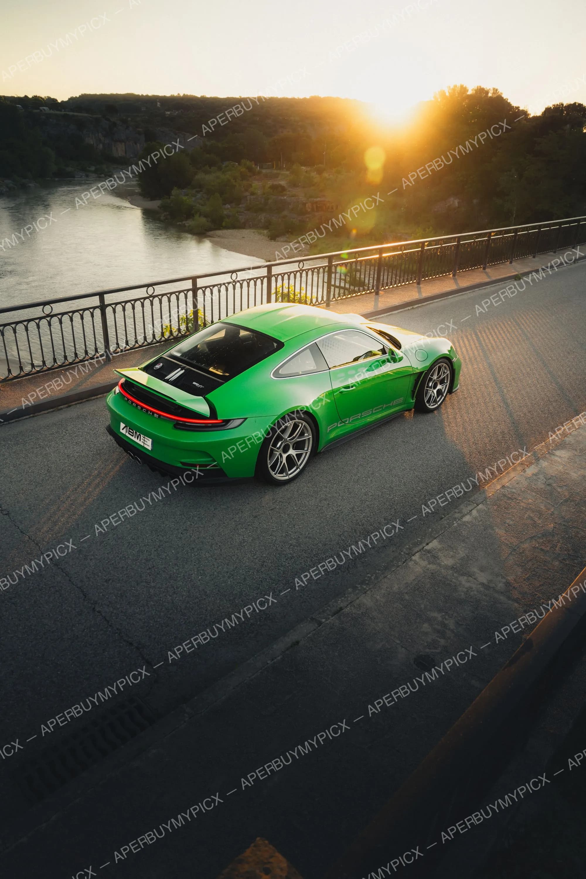 Porsche 911 (992) ST verte vue de trois-quarts arrière sur un pont au-dessus d'une rivière au coucher de soleil, jantes argentées et aileron déployé