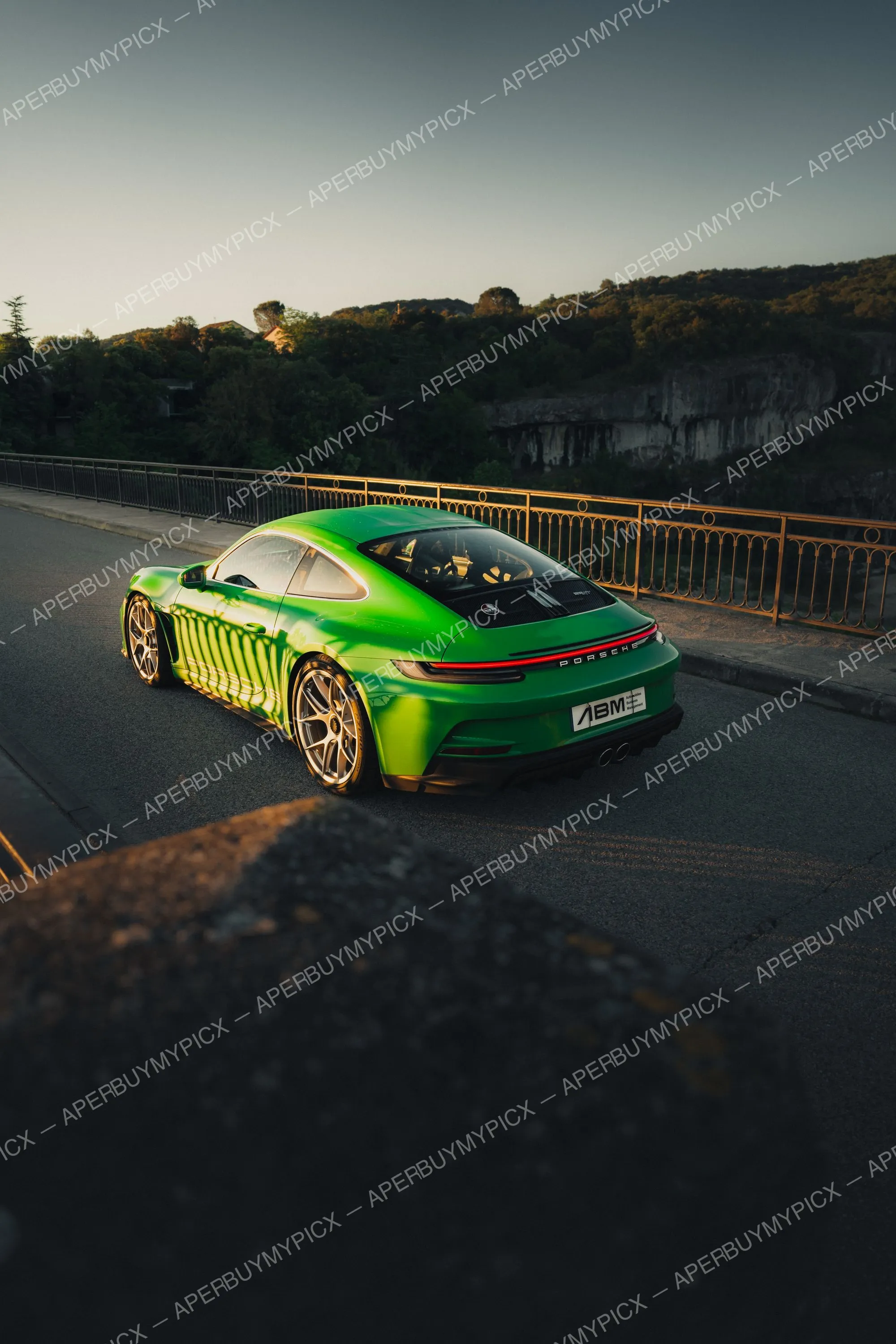 Porsche 911 992 ST verte vue de trois quarts arrière sur un pont bordé d'une rambarde dorée, jantes dorées, au coucher du soleil avec collines boisées et falaise en arrière-plan.