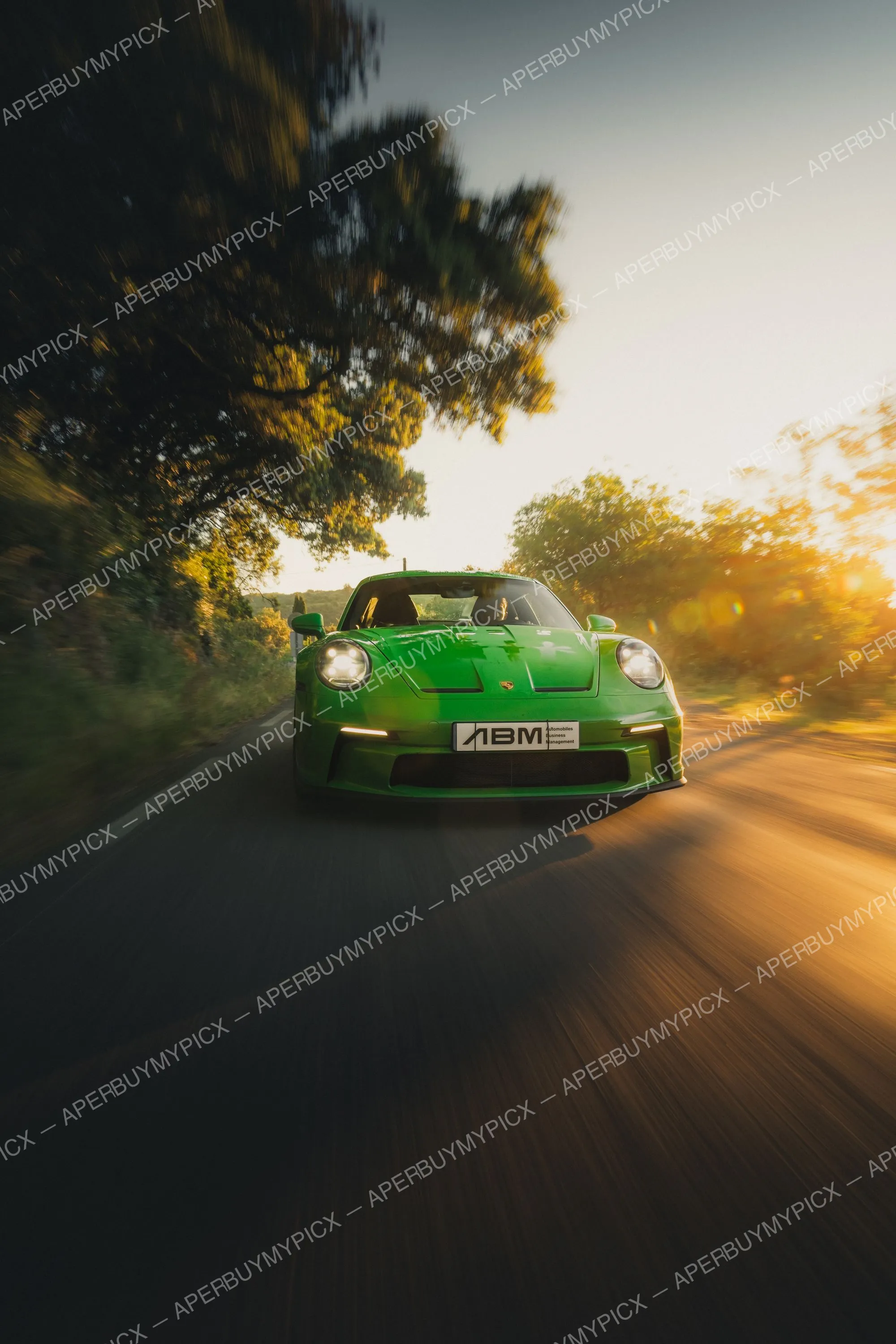 Porsche 911 992 ST verte roulant de face sur une route de campagne bordée d'arbres au coucher du soleil, phares allumés, plaque ABM, effet de vitesse.