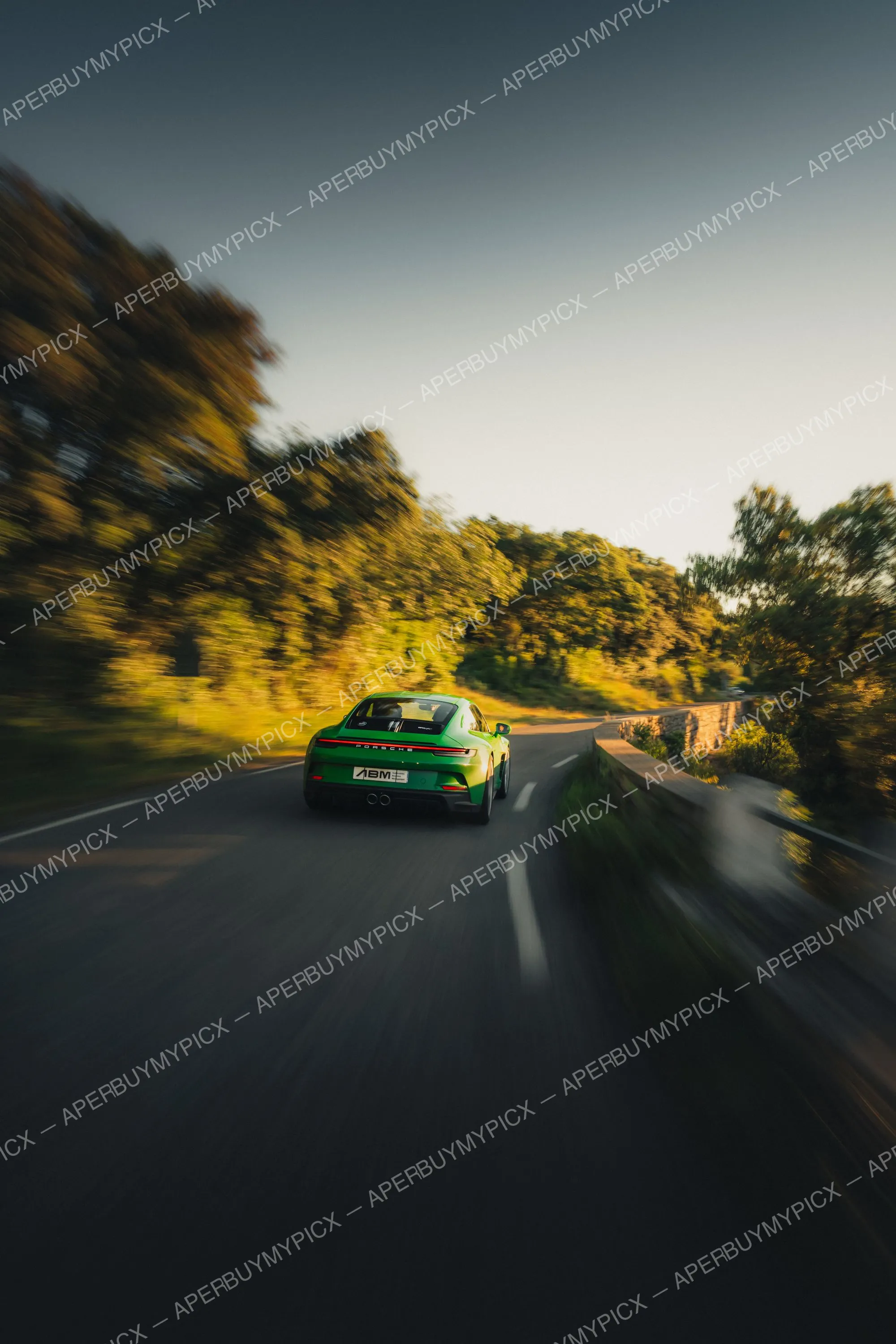 Porsche 911 992 ST verte vue de l'arrière roulant à vive allure sur une route de campagne bordée d'arbres au coucher du soleil, effet de flou de mouvement