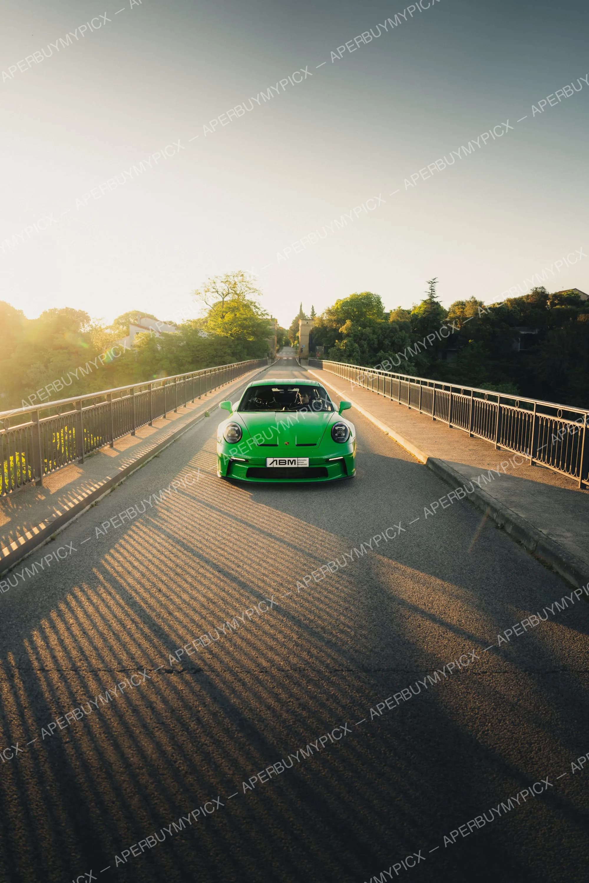 Porsche 911 GT3 verte vue de face, roulant sur un pont bordé de garde-corps au coucher du soleil, arbres et ciel dégagé en arrière-plan