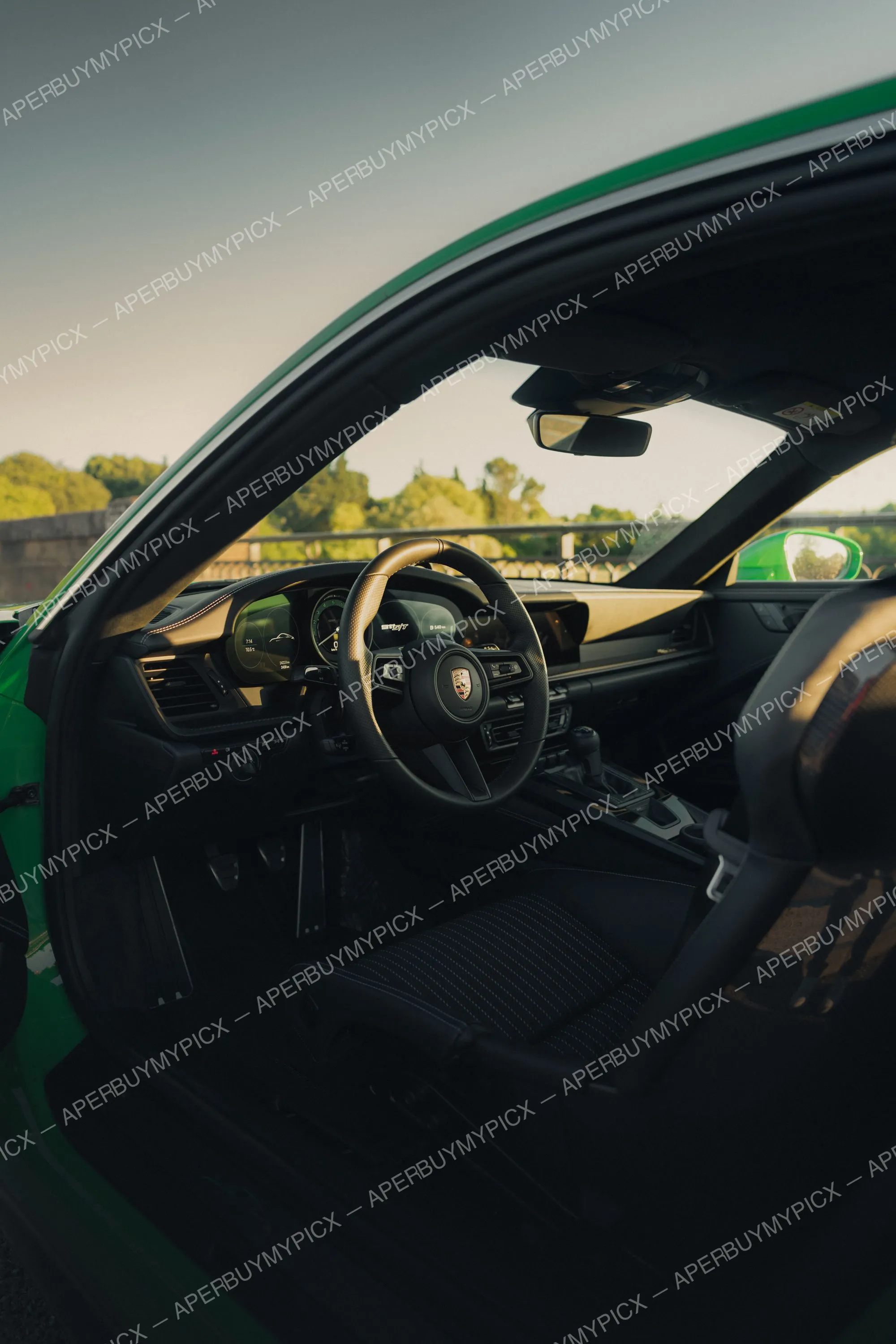 Intérieur d'une Porsche 911 (992) S/T verte vue depuis la portière ouverte, avec volant en cuir portant l'écusson Porsche, tableau de bord numérique et sièges sport, au coucher du soleil.