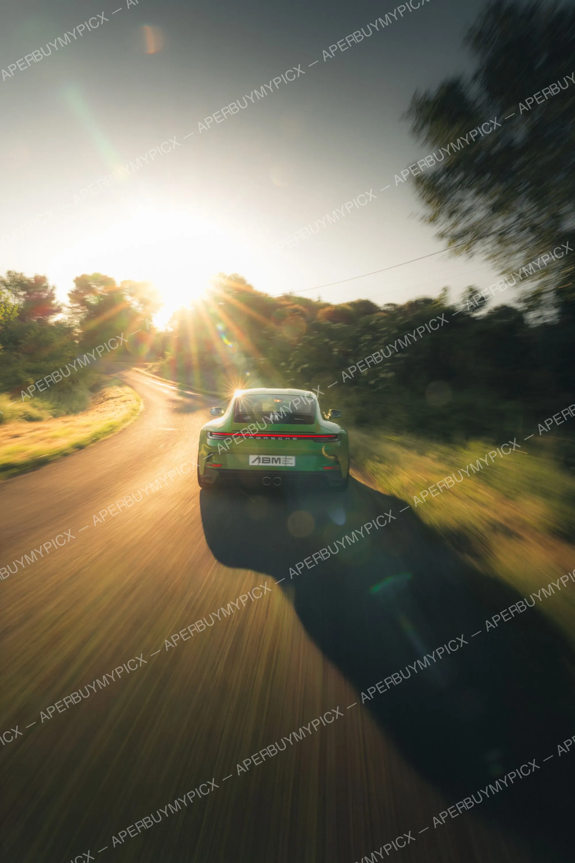 Porsche 911 992 ST verte vue de dos roulant sur une route de campagne au coucher du soleil, effet de vitesse et arbres flous