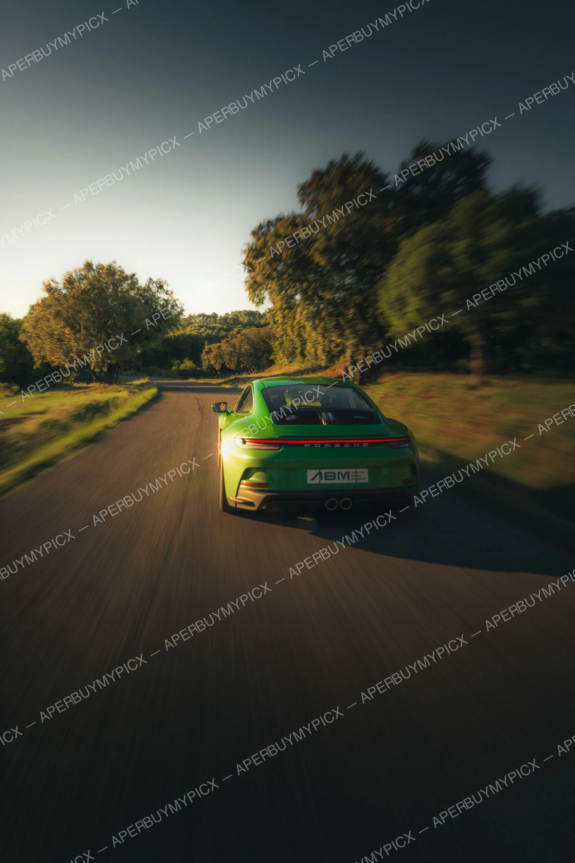 Porsche 911 992 ST verte vue de l'arrière roulant à vive allure sur une route de campagne bordée d'arbres au coucher du soleil, avec effet de flou de mouvement.