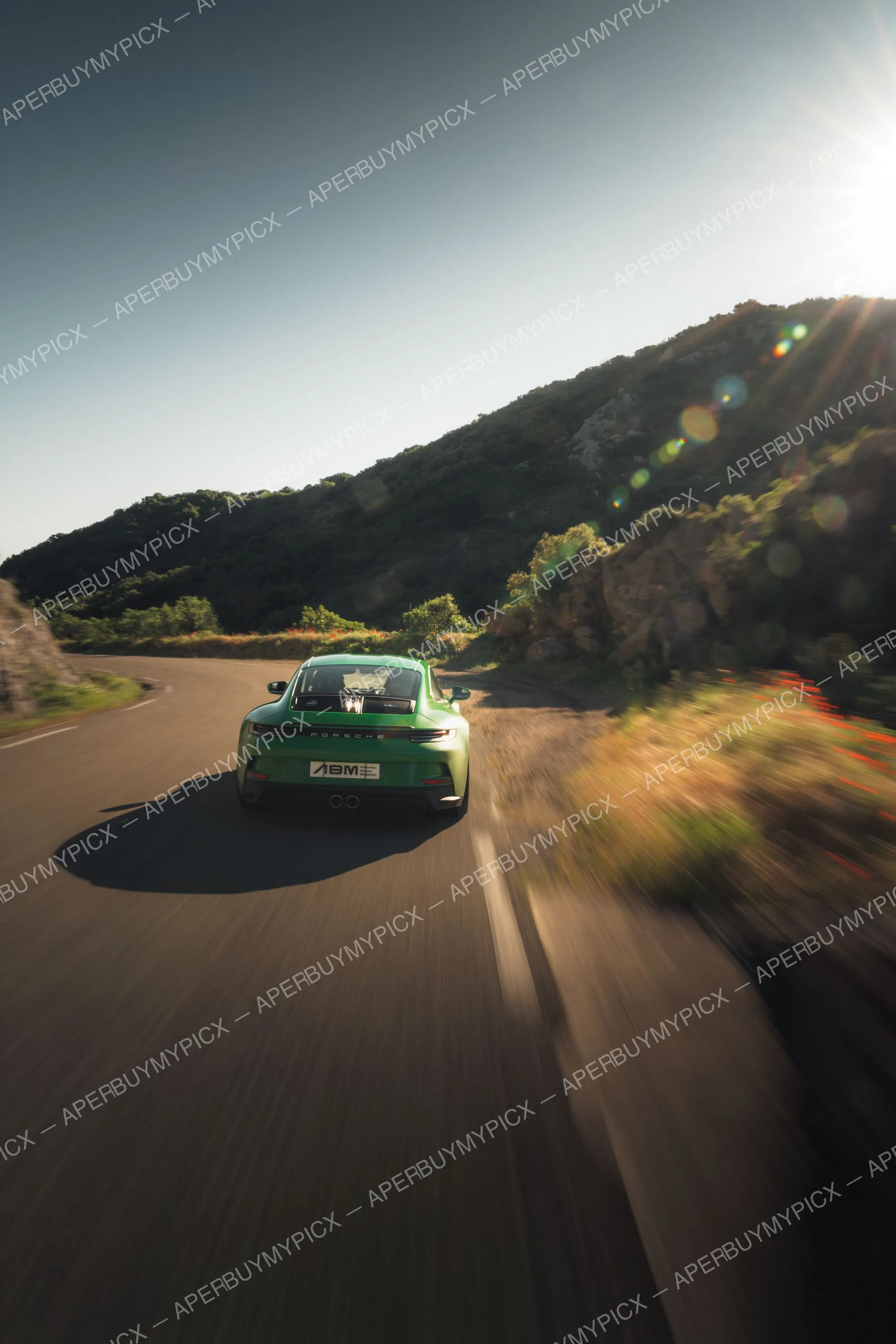 Porsche 911 992 ST verte vue de l'arrière roulant à vive allure sur une route de montagne sinueuse, avec effet de filé et soleil bas à l'horizon.