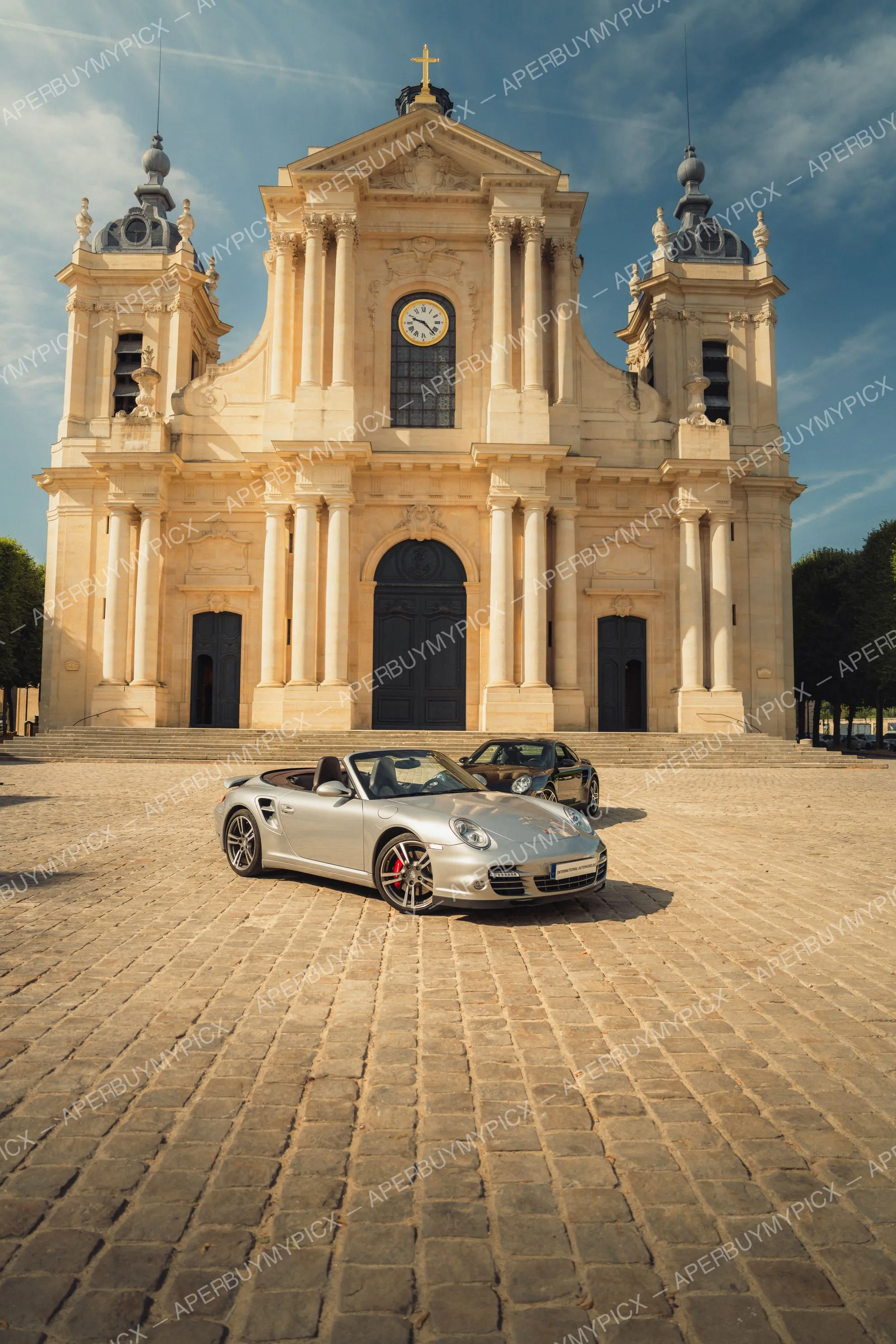 Porsche 911 Turbo Cabriolet argentée garée sur une place pavée devant une cathédrale baroque en pierre claire avec horloge et croix dorée, une Porsche noire à l'arrière-plan sous un ciel bleu.