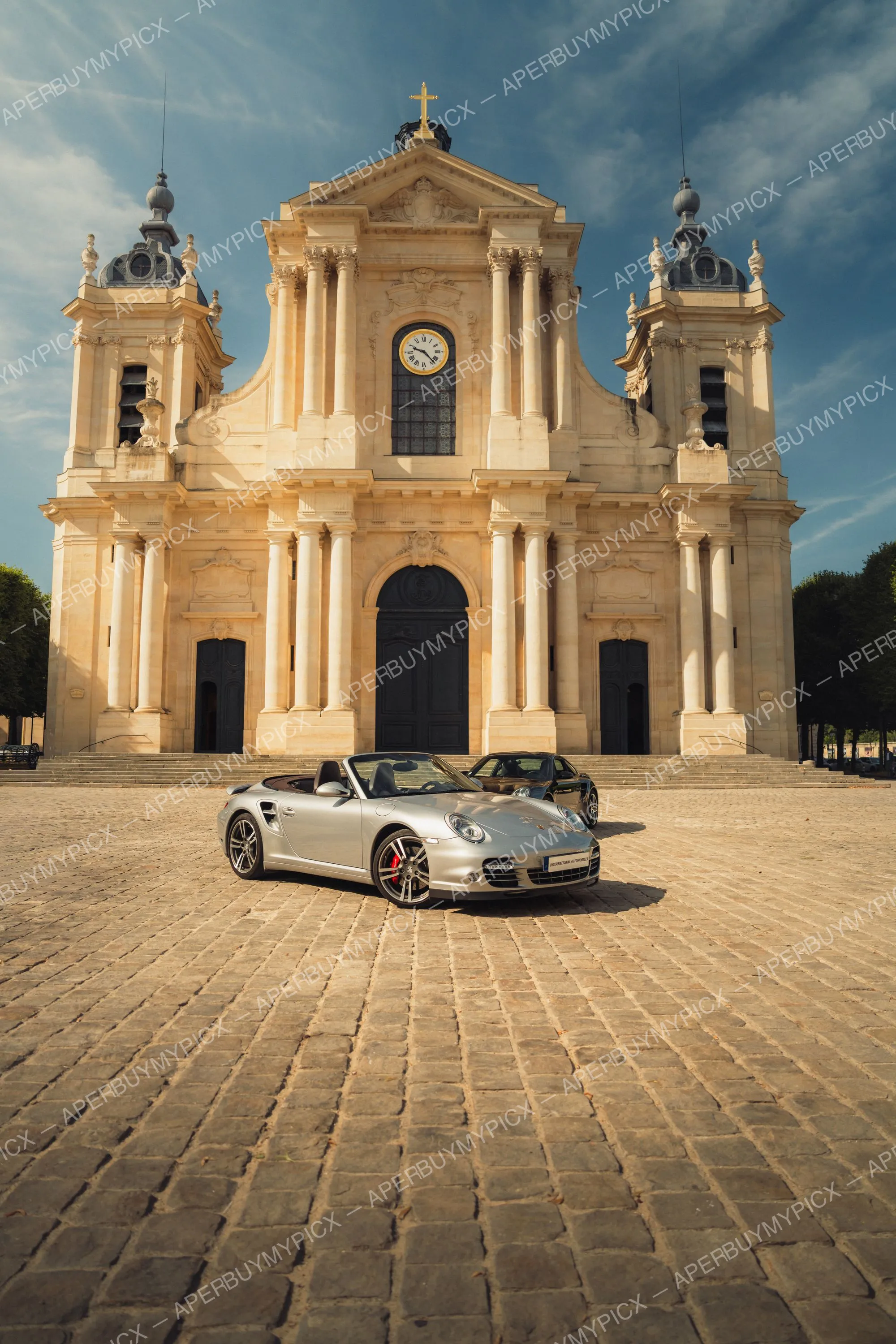 Porsche 911 Turbo cabriolet argentée garée sur une place pavée devant une cathédrale baroque à la lumière dorée, avec une seconde Porsche noire en arrière-plan