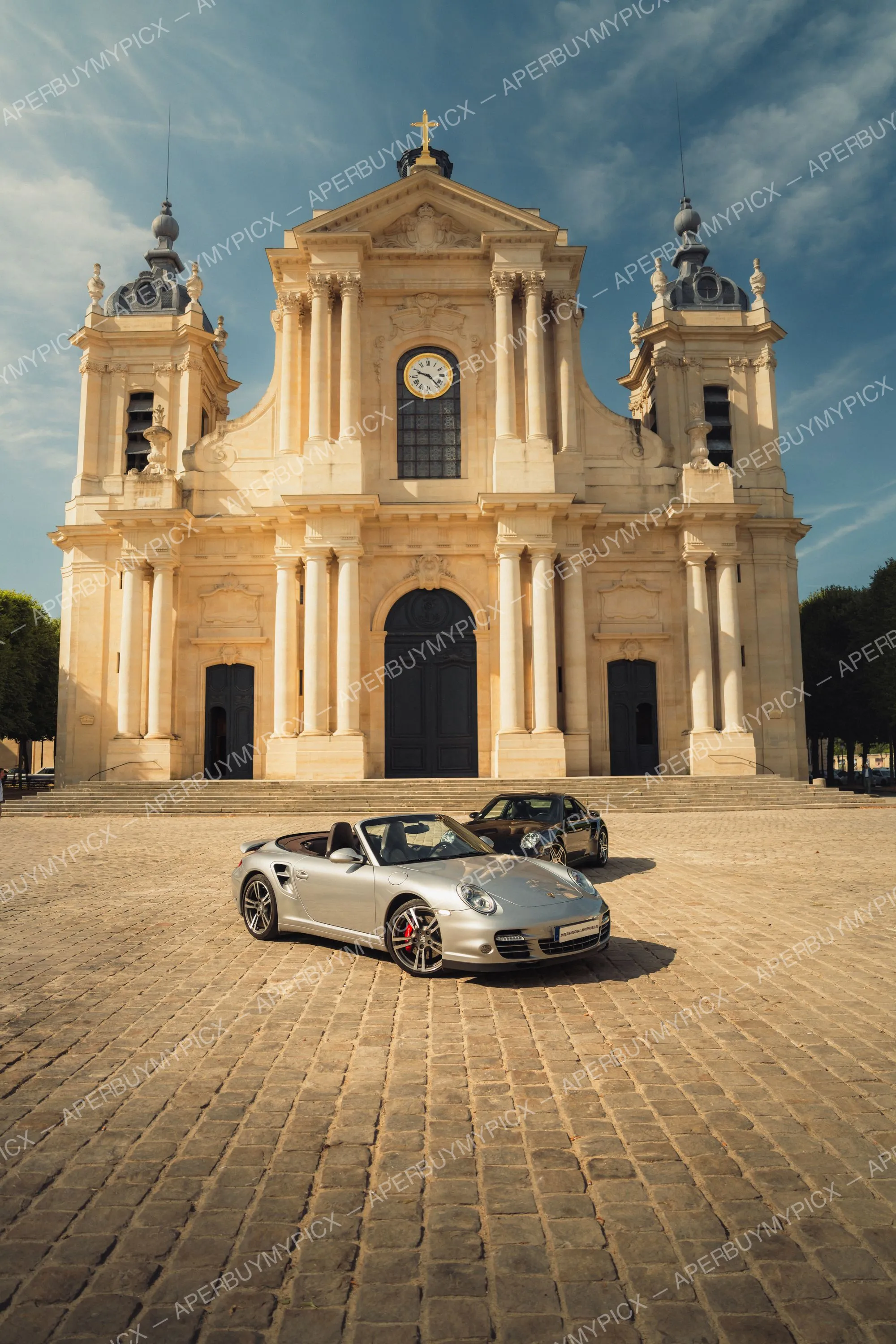 Porsche 911 Turbo Cabriolet argentée et coupé noir stationnées sur des pavés devant la façade d'une cathédrale baroque avec horloge et deux clochers, ciel bleu ensoleillé.