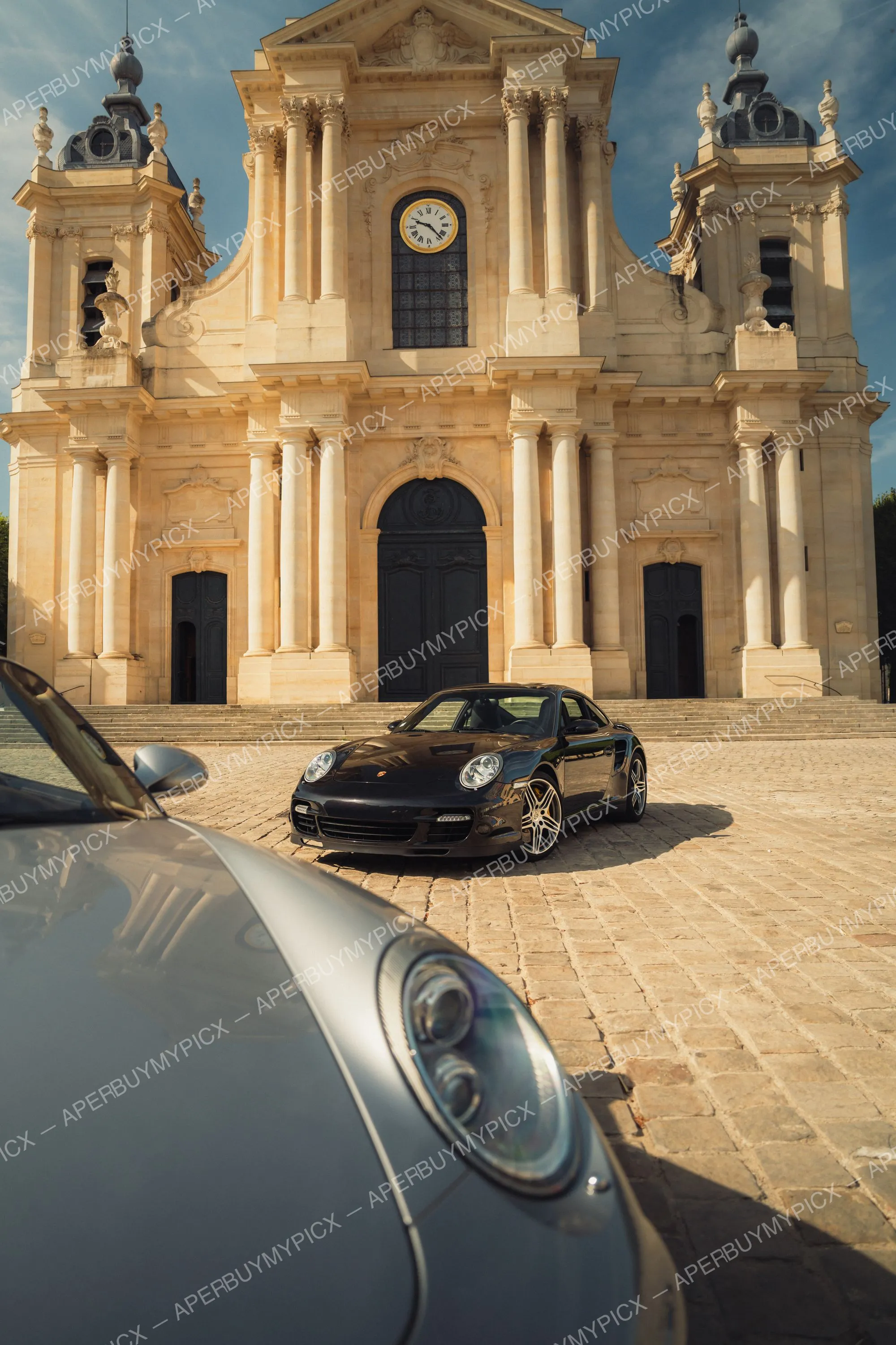 Porsche 911 Turbo noire garée sur une place pavée devant une cathédrale baroque avec horloge, à côté d'une Porsche grise au premier plan sous un ciel ensoleillé