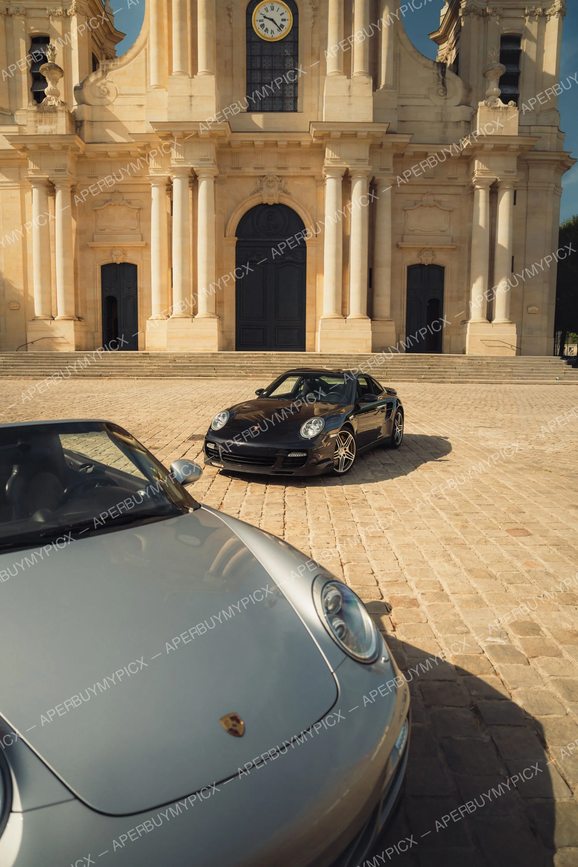 Porsche 911 Turbo noire stationnée sur des pavés devant une cathédrale baroque à horloge, avec l'avant d'une Porsche 911 grise au premier plan.
