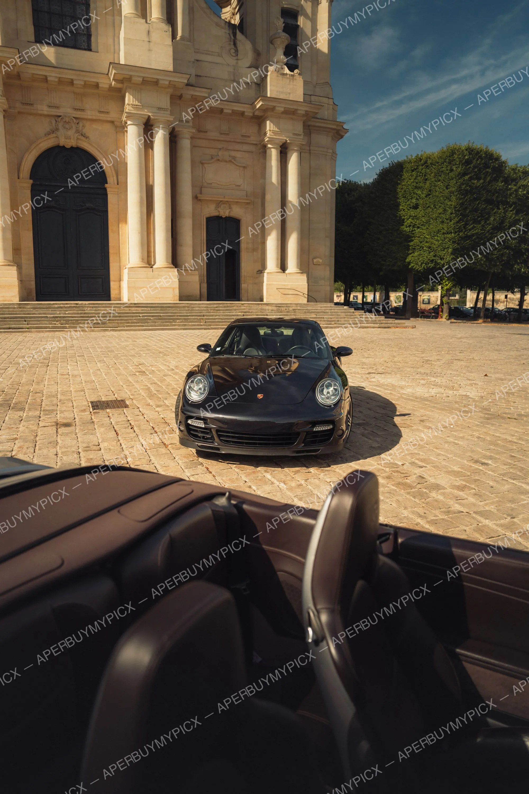 Porsche 911 Turbo noire garée sur une place pavée devant la façade en pierre d'une cathédrale baroque, vue de face depuis un cabriolet au coucher du soleil