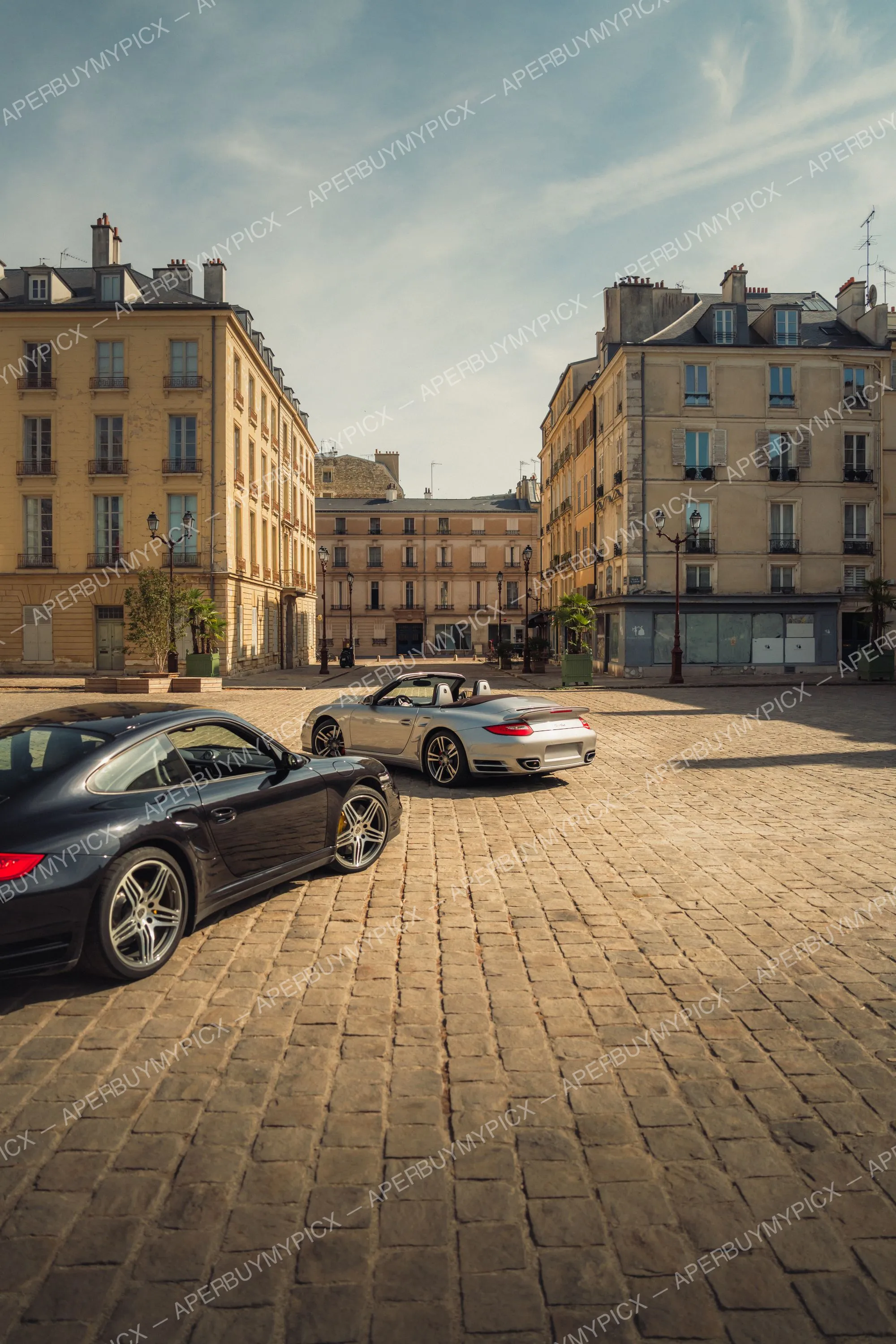 Une Porsche 911 Turbo noire coupé et une Porsche 911 Turbo cabriolet grise garées sur une place pavée entourée d'immeubles haussmanniens sous un ciel dégagé.