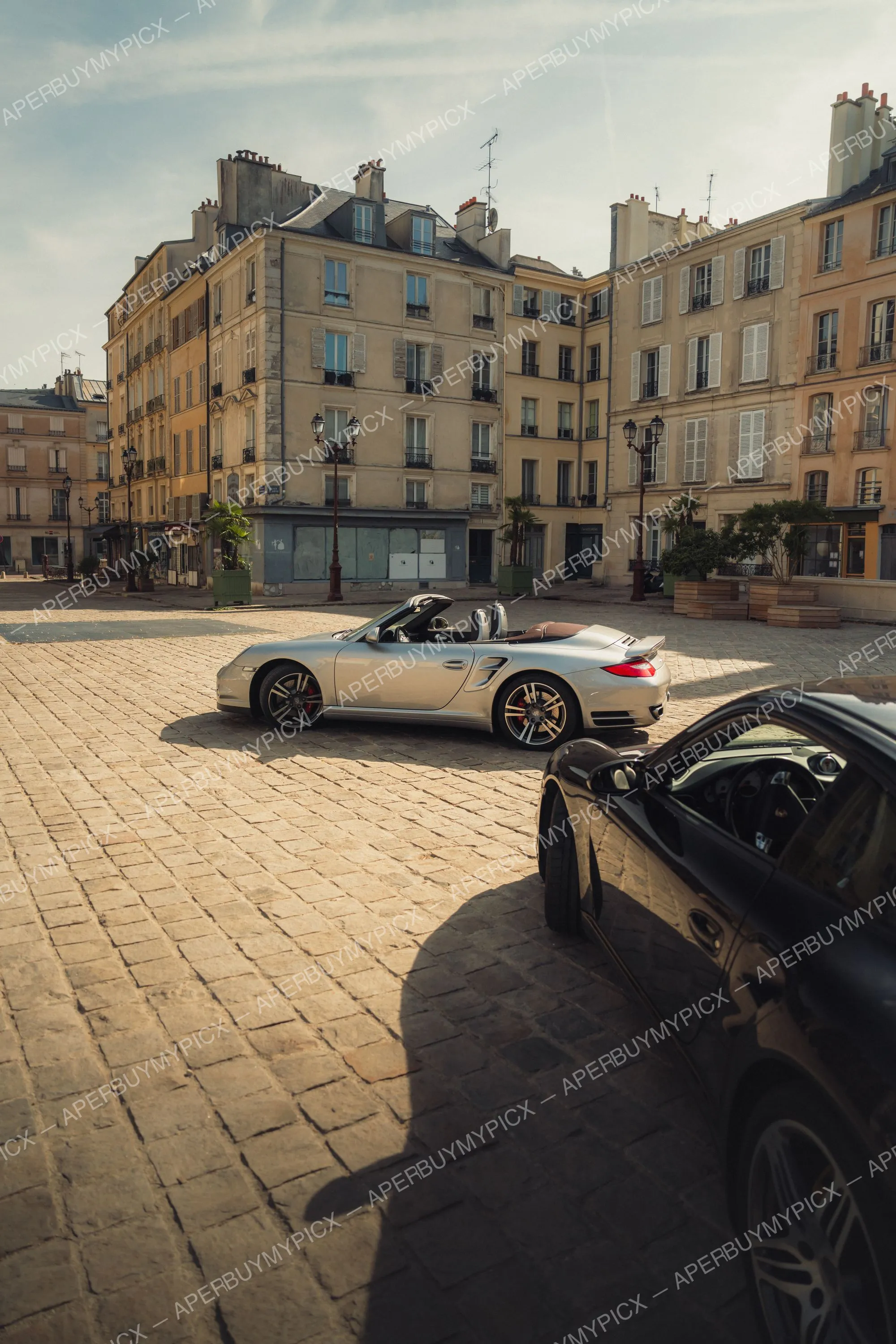 Porsche 911 Turbo cabriolet argentée garée sur une place pavée devant des immeubles haussmanniens, avec une Porsche noire au premier plan