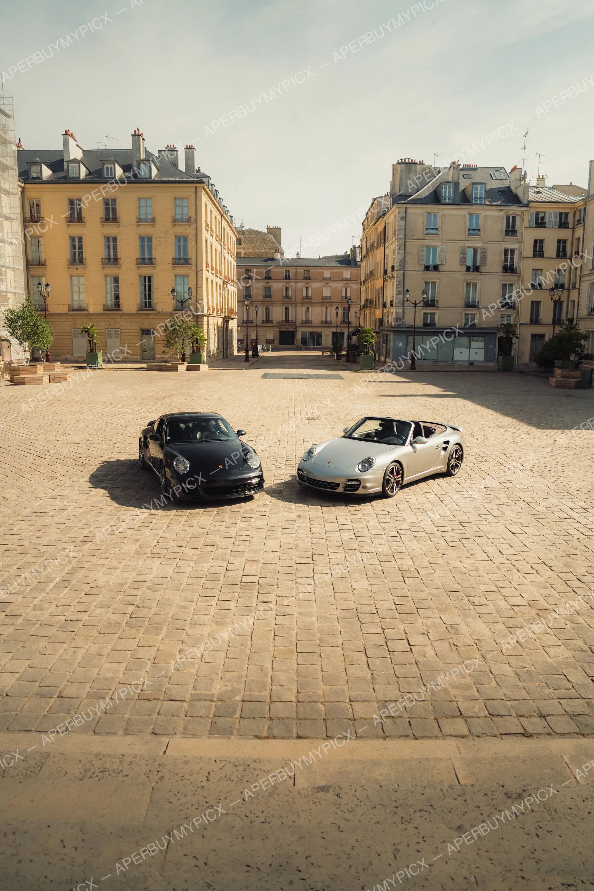 Deux Porsche 911 Turbo, une coupé noire et un cabriolet gris, garées face à face sur une place pavée parisienne bordée d'immeubles haussmanniens sous un ciel clair.