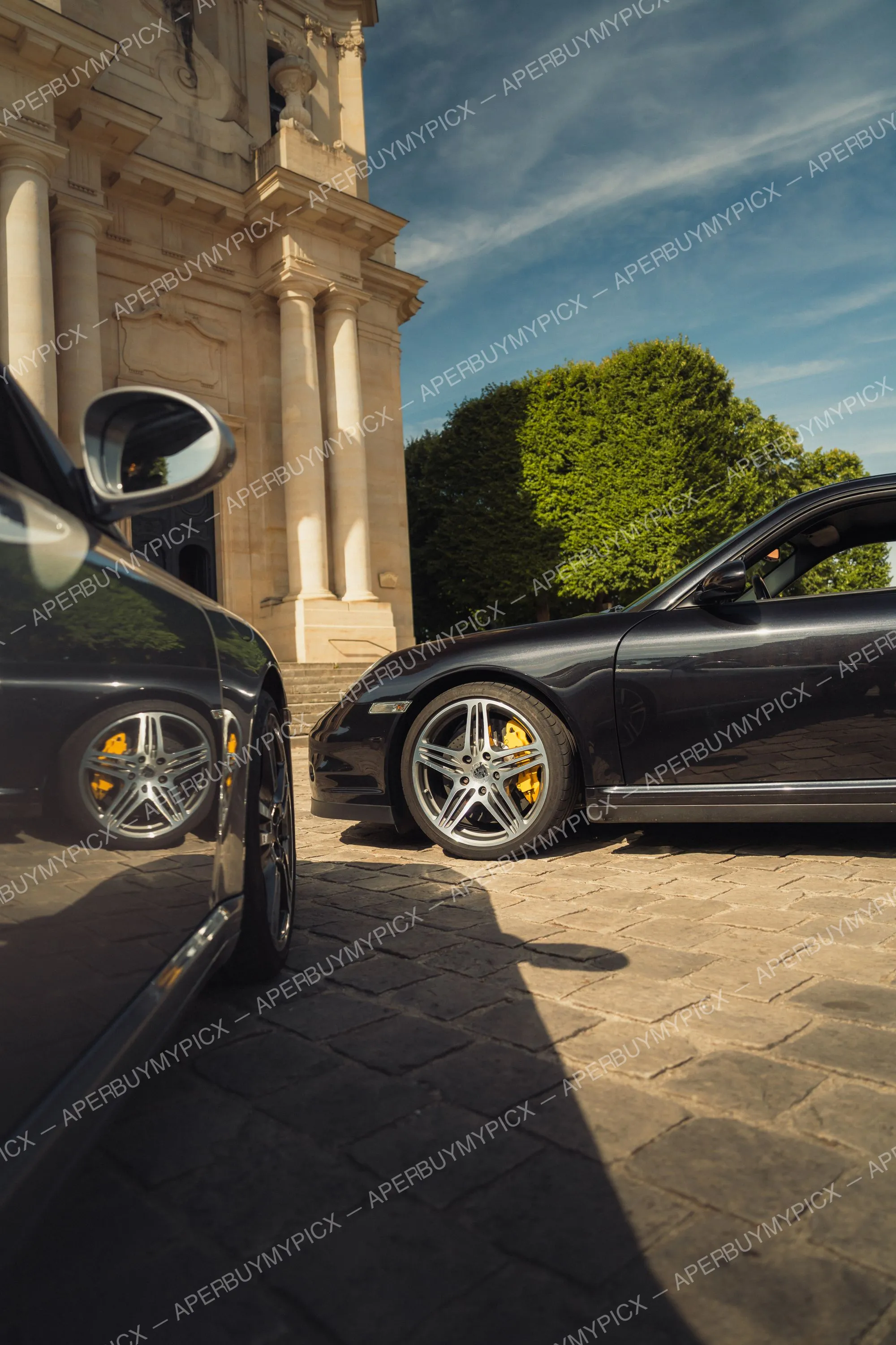Porsche 911 Turbo noire aux jantes chromées et étriers de frein jaunes, garée sur des pavés devant un bâtiment classique à colonnes, ciel bleu ensoleillé