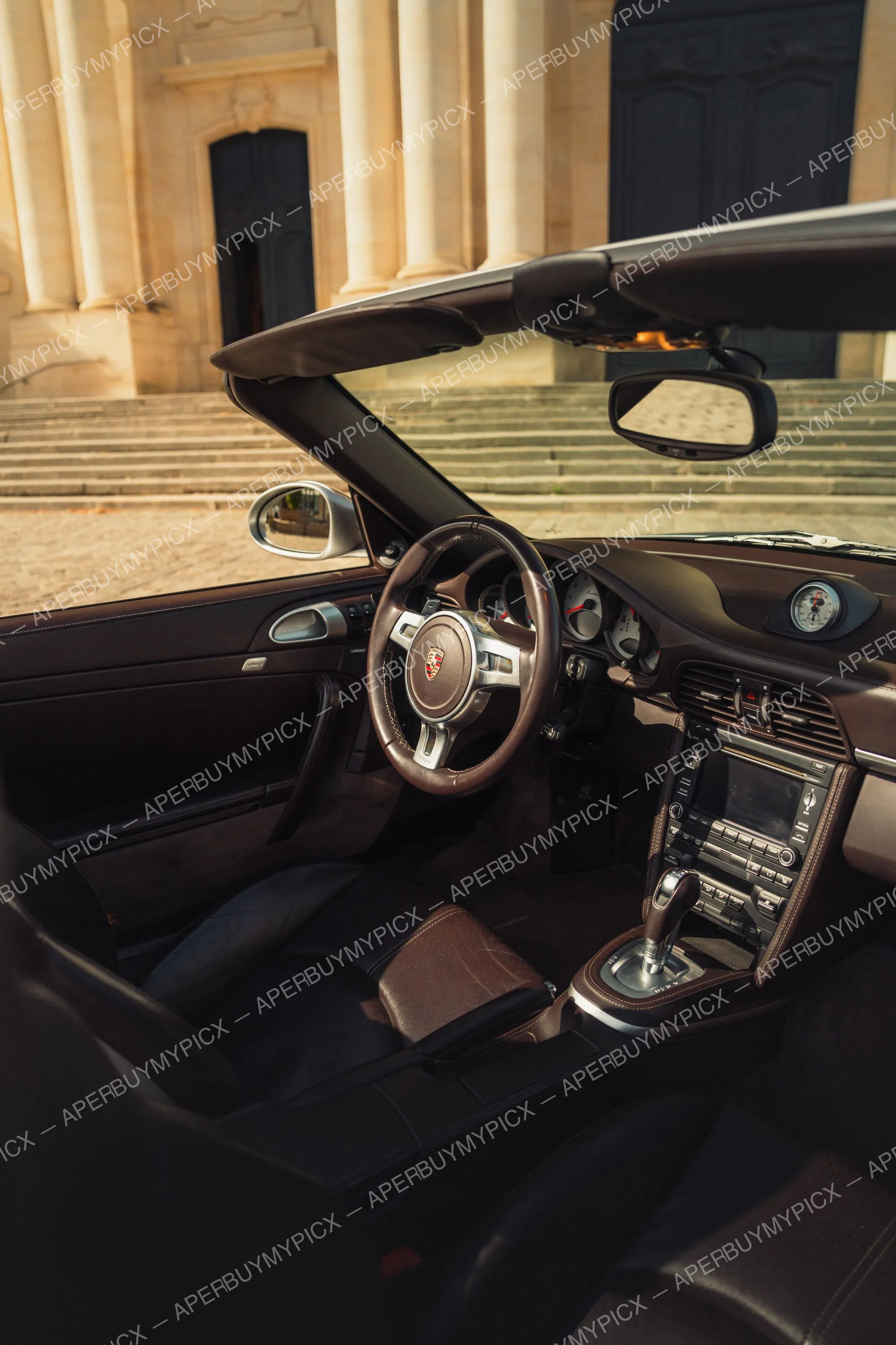 Intérieur d'une Porsche 911 Turbo cabriolet avec sièges et volant en cuir marron, tableau de bord et console centrale, garée devant un bâtiment en pierre.